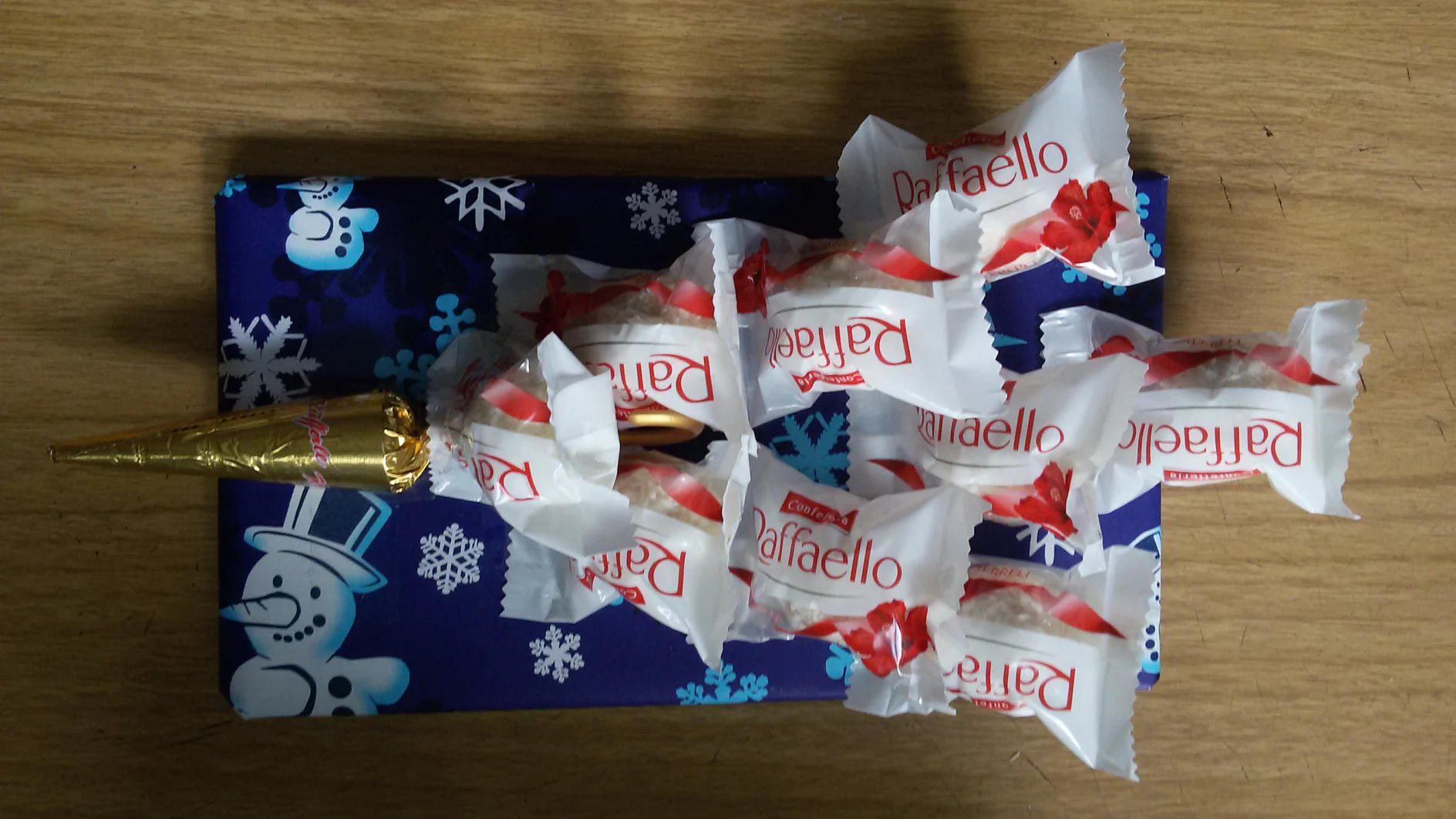 Ein dekoratives Arrangement bestehend aus einer kleinen, in Gold eingewickelten Schokoladentüte, gekrönt mit einzeln verpackten Raffaello-Bonbons, die auf einem rechteckigen Objekt ruht, das in dunkelblaues Papier mit Schneemannmuster eingewickelt und mit Tesaband befestigt ist. Auf einer Holzoberfläche platziert. (This text has been generated by AI)
