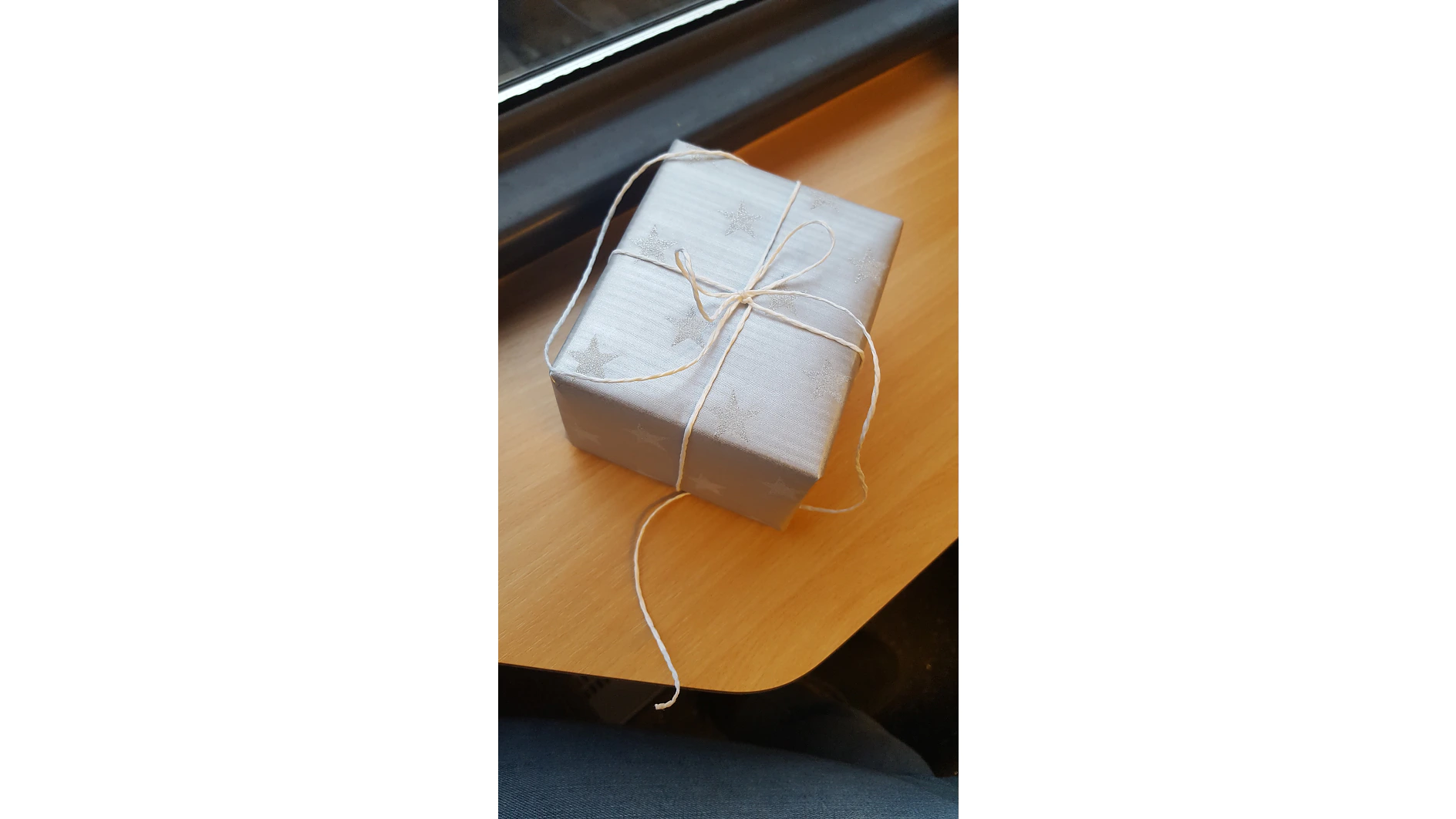 Auf einer Holzfläche steht eine kleine, quadratische Geschenkschachtel, eingewickelt in silbernes Papier mit Sternenmuster, befestigt mit Tesa-Klebeband und einer weißen Schleife. (This text has been generated by AI)