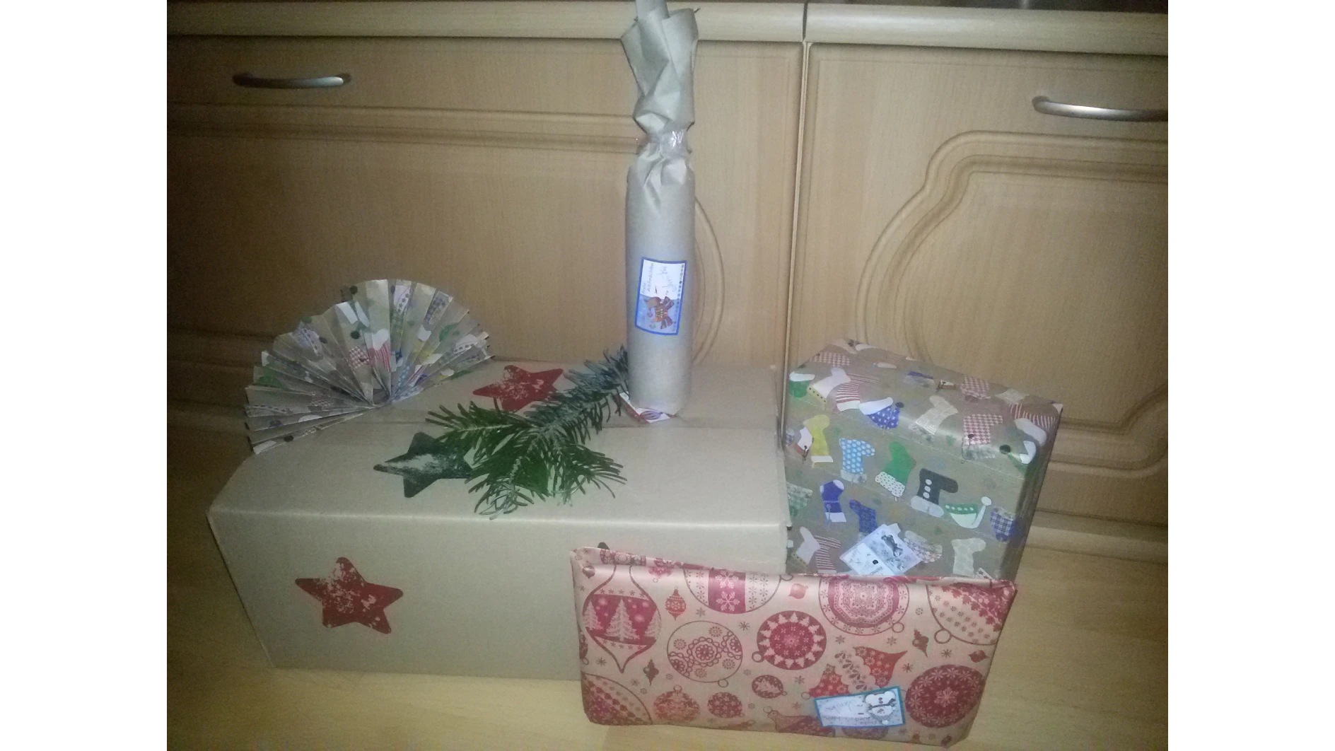 Mehrere verpackte Geschenke liegen auf einem Holzboden vor einem Schrank. Das größte Geschenk ist in schlichtes Papier mit roten Sternen eingewickelt und mit Tesa-Klebeband befestigt. Andere Geschenke haben buntes, festliches Papier, alles nahtlos mit Tesa-Klebeband zusammengehalten. Ein dekorativer Papierfächer und ein kleiner Zweig begleiten das Arrangement. (This text has been generated by AI)