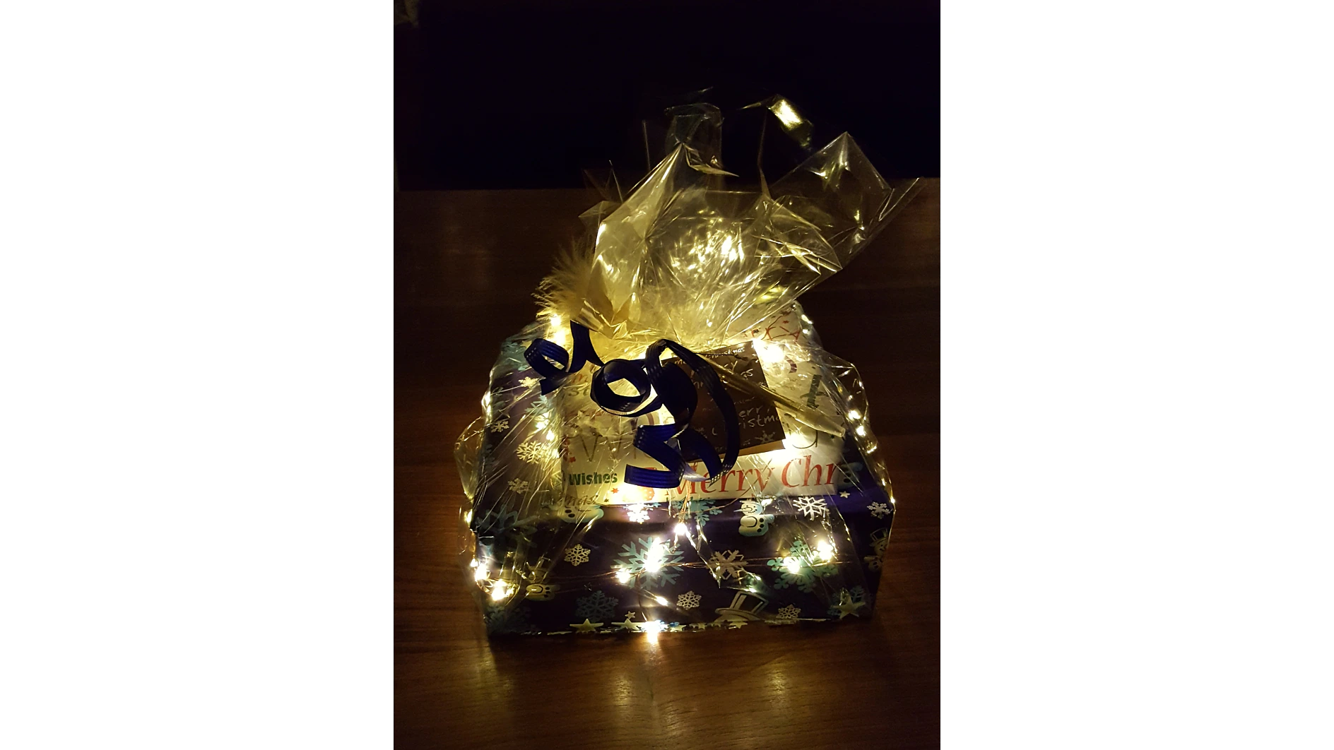 Ein Geschenk, eingewickelt in festliches Papier, geschmückt mit Schneeflocken und den Worten „Frohe Weihnachten“. Das Paket ist mit durchsichtigem Kunststoff überzogen, mit einer goldenen Schleife und einem blauen Band verziert, alles zusammengehalten mit Tesa-Klebeband. Kleine Lichterketten erhellen die Szene. (This text has been generated by AI)