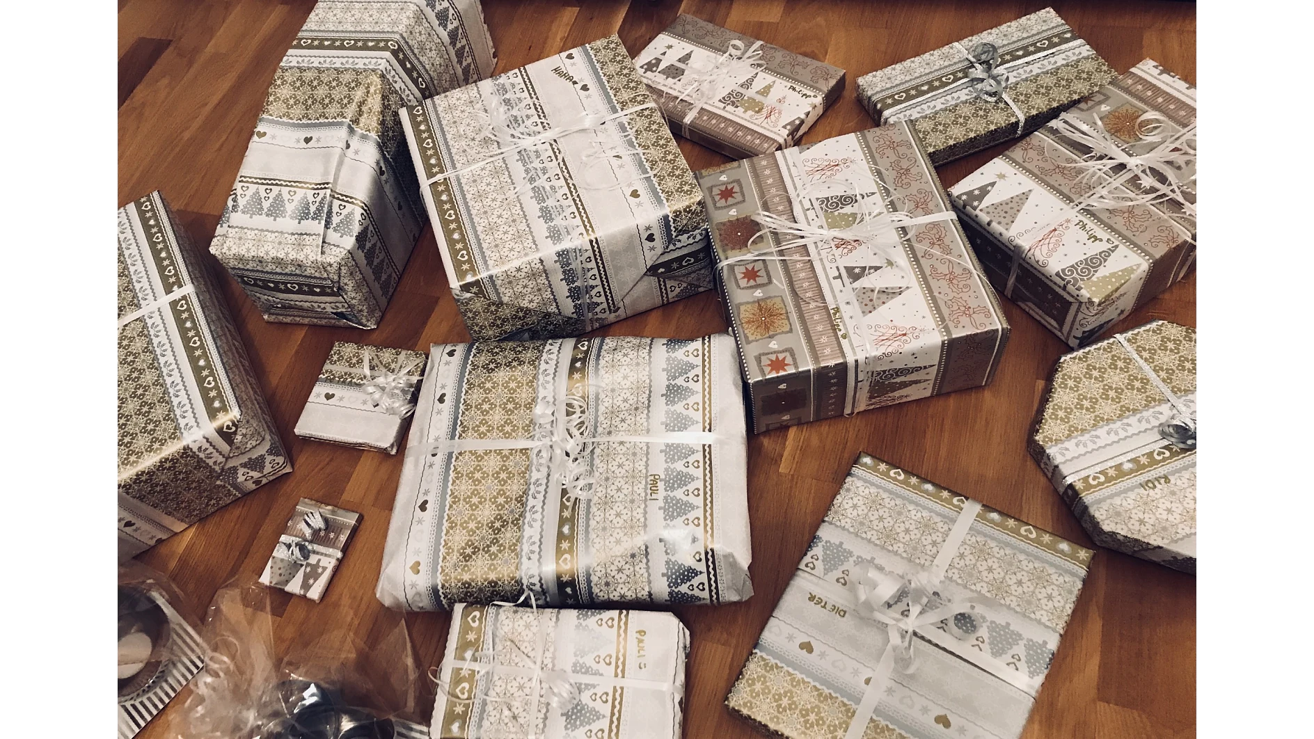 Eine Sammlung von Geschenken in verschiedenen Größen und Formen, verpackt in festliches, gemustertes Papier mit Tesa-Klebeband, Schleifen und Bändern. Sie sind auf einem Holzboden angeordnet. Das Geschenkpapier weist Designs mit Bäumen, Sternen und geometrischen Mustern auf. (This text has been generated by AI)