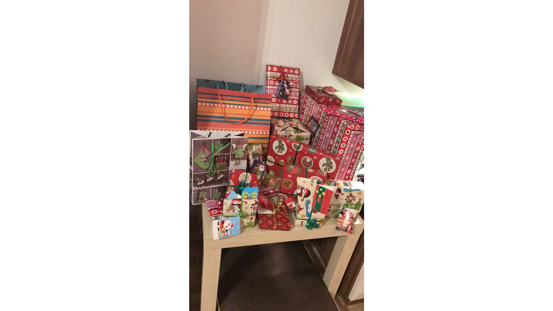 Ein Tisch, der mit einer Vielzahl verpackter Weihnachtsgeschenke bedeckt ist, die alle mit Tesa-Klebeband befestigt sind. Die Geschenke sind in festliches Papier mit Mustern wie Schneemännern, Rentieren und Ornamenten eingewickelt. Eine Mischung aus Kartons und Taschen ist übereinander gestapelt, wobei eine nahe gelegene Wand und ein Teil eines Holzschranks sichtbar sind. (This text has been generated by AI)