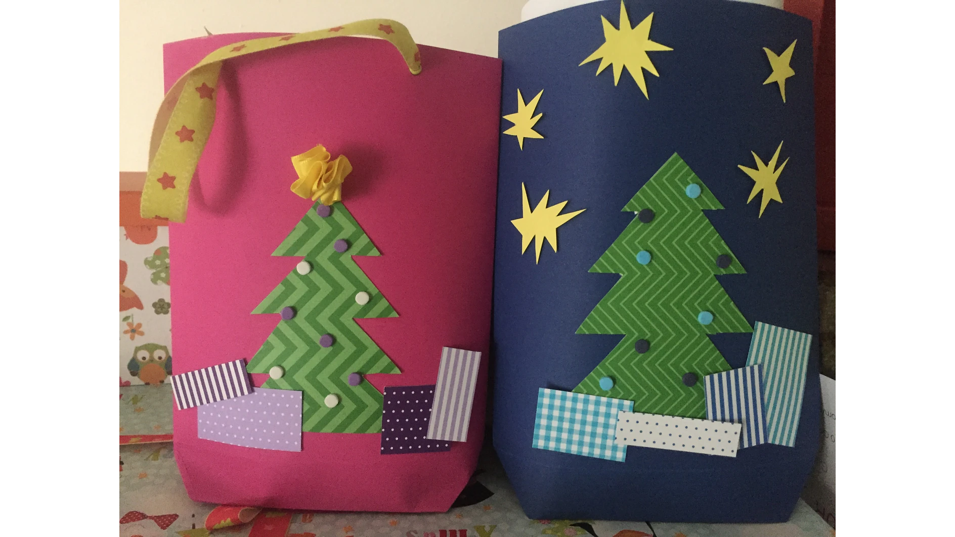 Es werden zwei dekorierte Papiertüten gezeigt. Die rosa Tüte links hat einen Griff und zeigt unten einen grünen Zickzack-Weihnachtsbaum mit violetten Punkten und bunten Rechtecken. Die blaue Tüte rechts hat gelbe Sternformen und ein ähnliches Baumdesign, alles zusammengehalten mit Tesa-Klebeband. (This text has been generated by AI)