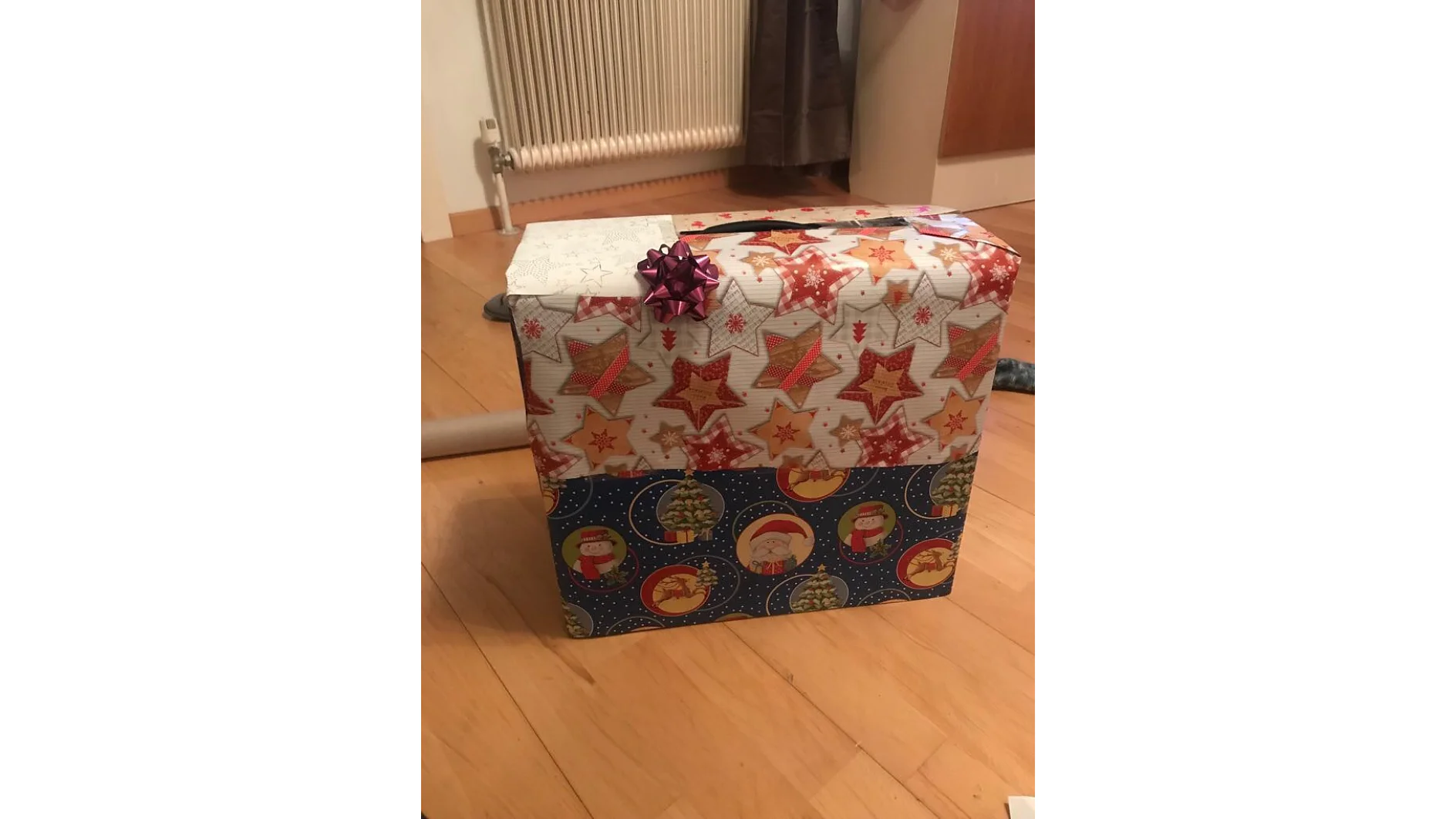 Auf einem Holzboden steht ein großes, quadratisches Geschenk, das in festliches Papier mit Sternen und Weihnachtsmotiven eingewickelt ist. Darauf ist eine kleine lila Schleife, die mit Tesaband befestigt ist. Im Hintergrund sind ein Heizkörper und ein Teil einer Tür zu sehen. (This text has been generated by AI)