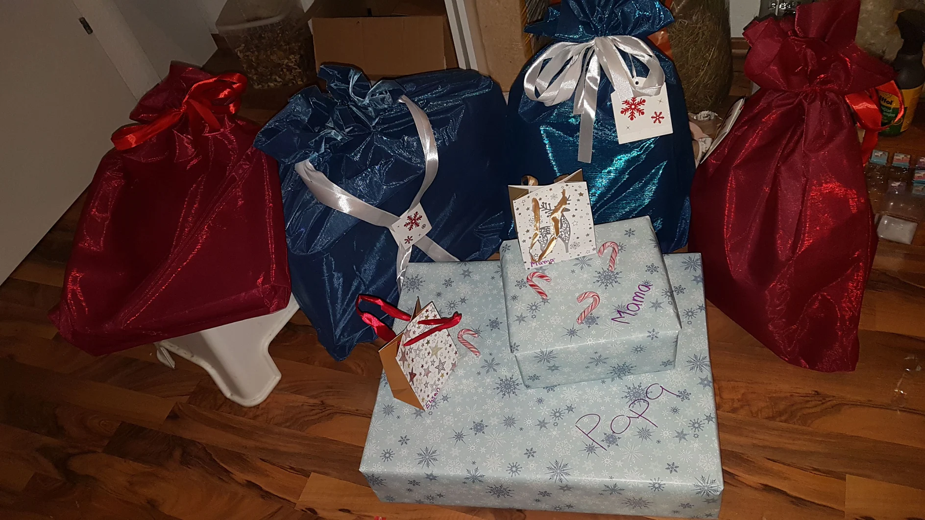 Mehrere verpackte Geschenke sind auf einem Holzboden angeordnet. Die Pakete sind in glänzenden Tüten und Kartons, hauptsächlich in Rot, Blau und Weiß mit festlichen Mustern. Einige haben Namensschilder, die mit Tesa-Klebeband befestigt sind und handgeschriebene Namen und Zuckerstangendekorationen aufweisen. (This text has been generated by AI)
