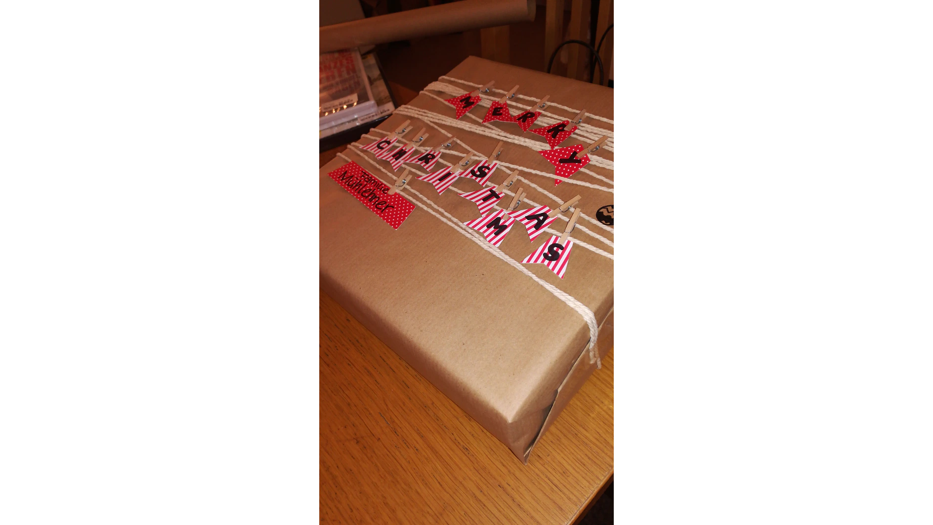 Ein in braunes Papier eingewickeltes Geschenk mit kleinen rot-weiß gestreiften Papierfähnchen, die mit Mini-Wäscheklammern befestigt sind. Schwarze Buchstaben auf den Fähnchen ergeben „Weihnachten“. Auf einem roten Stoffstück mit Punkten steht „Frohe Weihnachten“. Das Geschenk liegt auf einem Holztisch und ist mit Tesa-Klebeband sicher eingewickelt. (This text has been generated by AI)