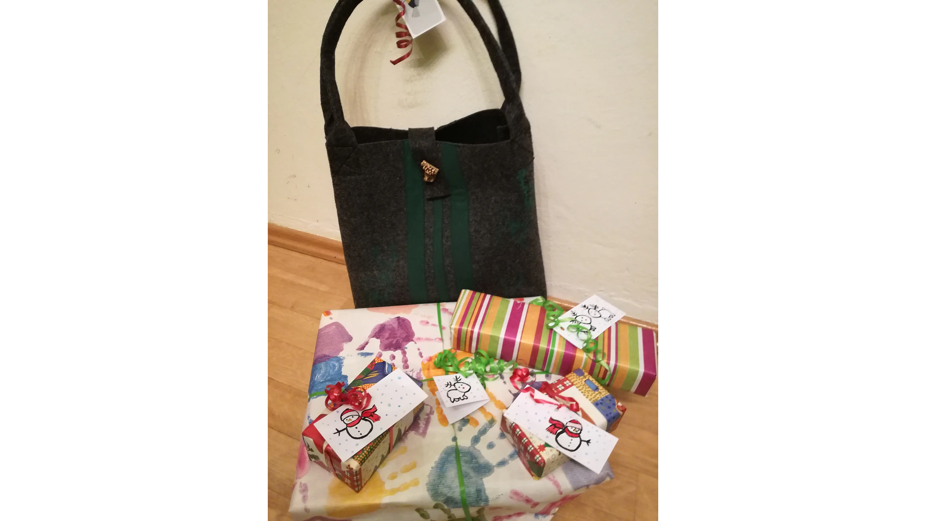 An einer Wand steht eine Filzhandtasche mit dunklen Riemen. Davor liegen vier in buntes Papier eingewickelte Geschenke, jedes mit Schleifen und kleinen Karten mit Schneemann-Motiven geschmückt. Die Geschenke sind mit Tesa-Klebeband zusammengehalten und auf einem Holzboden platziert. (This text has been generated by AI)