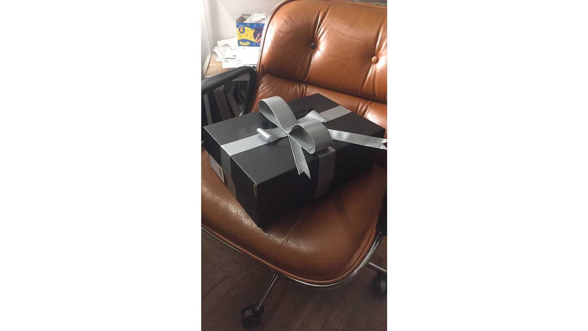 Eine große schwarze Geschenkbox mit silberner Schleife steht auf einem braunen Ledersessel. Im Hintergrund steht ein Regal mit einer Schachtel Tesa-Klebeband und anderen Gegenständen. Der Boden ist aus Holz. (This text has been generated by AI)