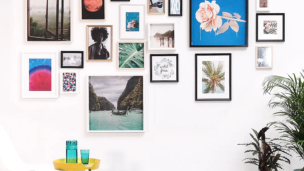 Certainly! Here is the revised text: A white wall displays a diverse collection of framed art, including nature photographs, abstract designs, and a rose painting. Below, a yellow table with two teal glasses is beside a white chair. A dark rug and two potted plants complete the scene. All artwork is meticulously hung using tesa tape for a secure display without any damage to the wall. (This text has been generated by AI)