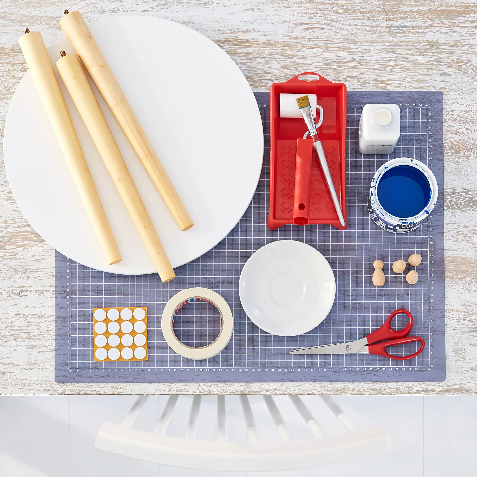 Tabletop with craft materials: three wooden dowels, white round panel, paint tray with a brush, open blue paint can, white bottle, adhesive dots, tesa tape, white dish, red scissors, and small wooden pegs on a cutting mat. (This text has been generated by AI)