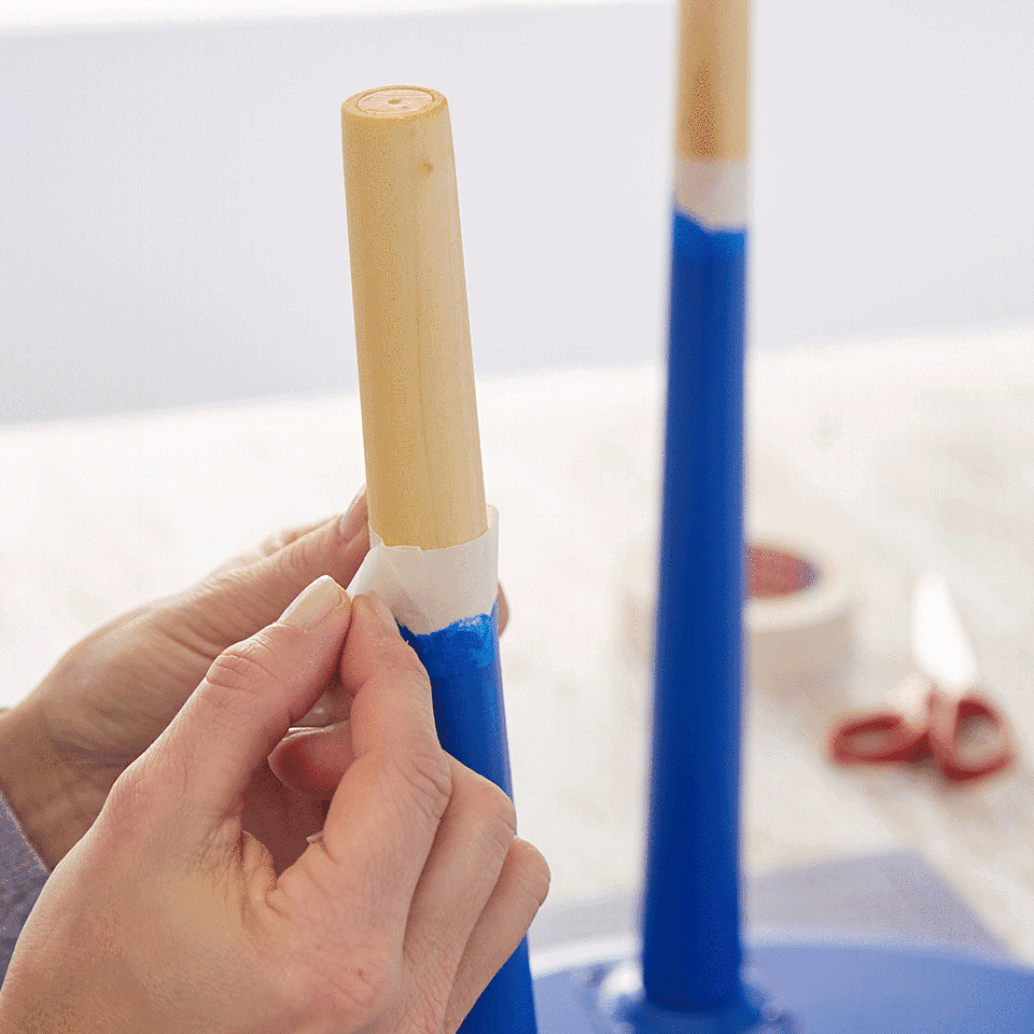 Hands are peeling off tesa tape from a wooden table leg painted blue at the bottom. Another similarly painted leg is visible in the background. Scissors and a roll of tesa tape are on the surface nearby. (This text has been generated by AI)
