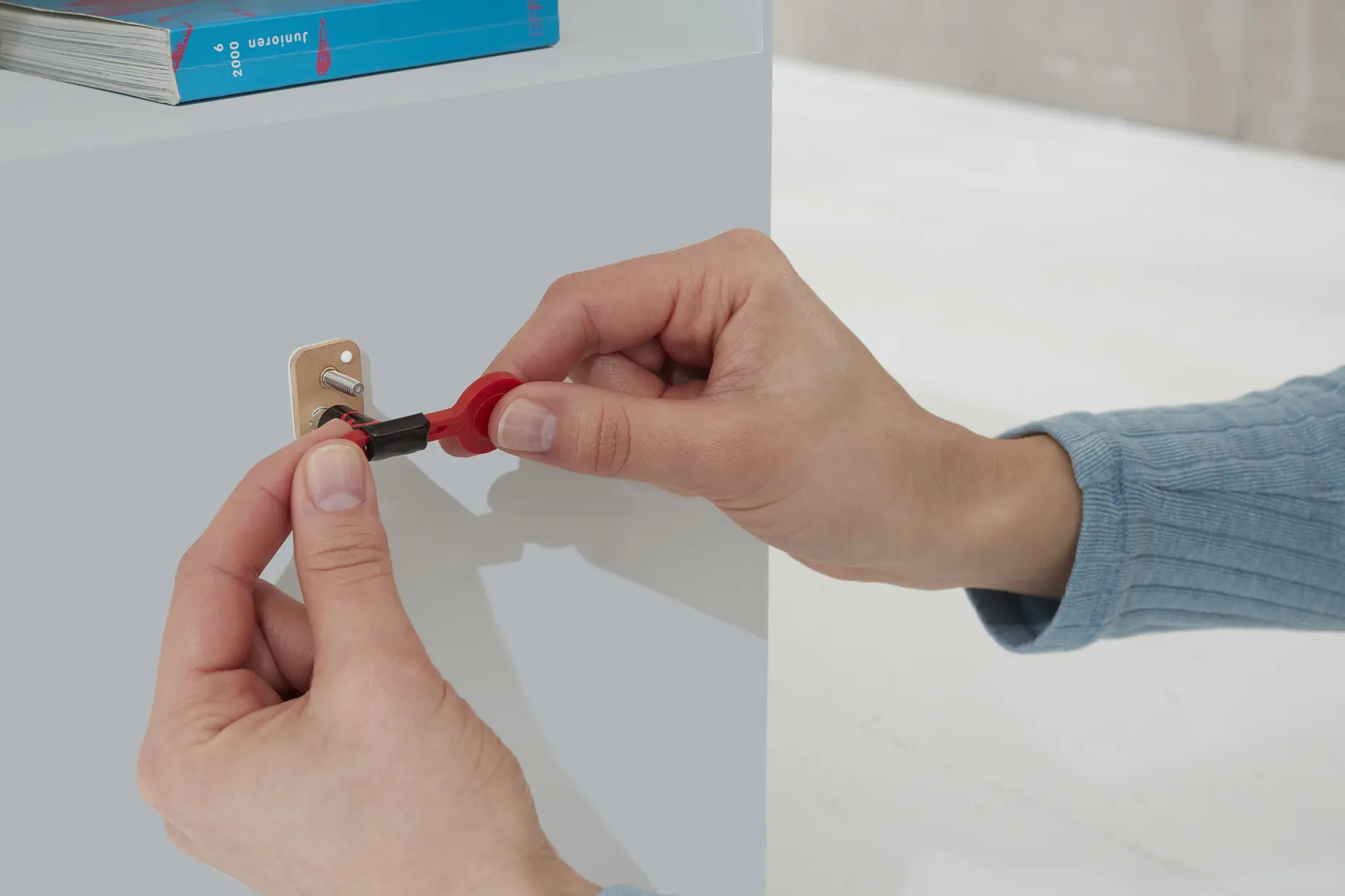 A person wearing a blue long-sleeve shirt is inserting a small screwdriver into a brass hinge attached to a light gray cabinet using tesa tape. A stack of blue books is visible on top of the cabinet. (This text has been generated by AI)