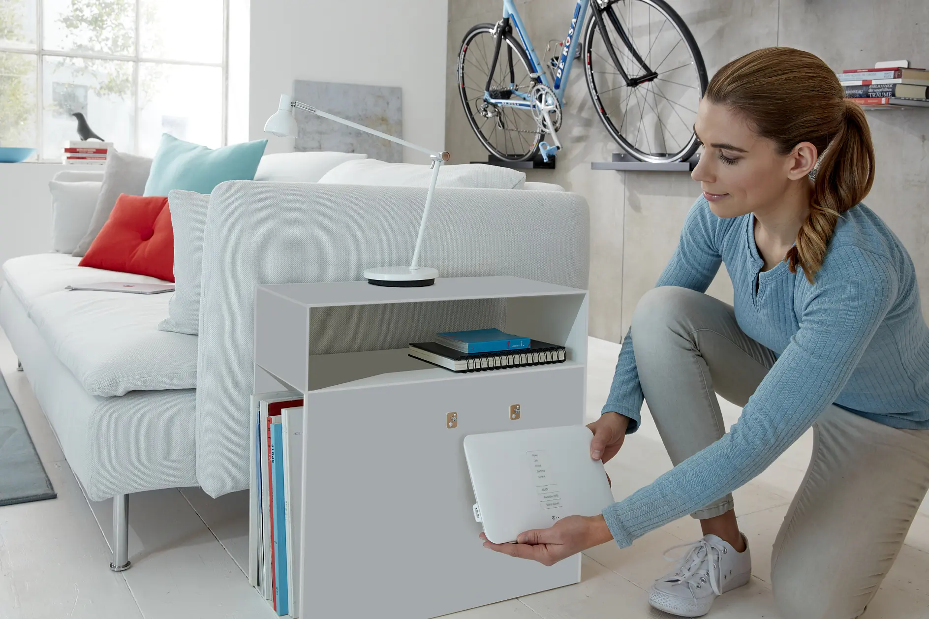A woman in a light blue sweater kneels beside a white cabinet with open shelving, placing or removing a Wi-Fi router. A sofa, books, a desk lamp, and a mounted bicycle are visible in the modern room. (This text has been generated by AI)