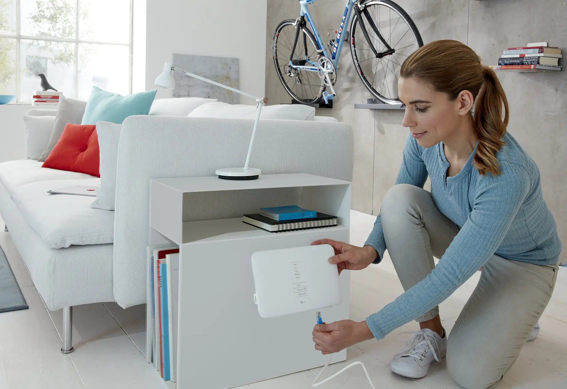 A woman is kneeling beside a white bookshelf in a modern living room, connecting a white electronic device with tesa tape. The room features a white sofa, a wall-mounted bicycle, a small lamp, and books. The space is bright with natural light from a window. (This text has been generated by AI)