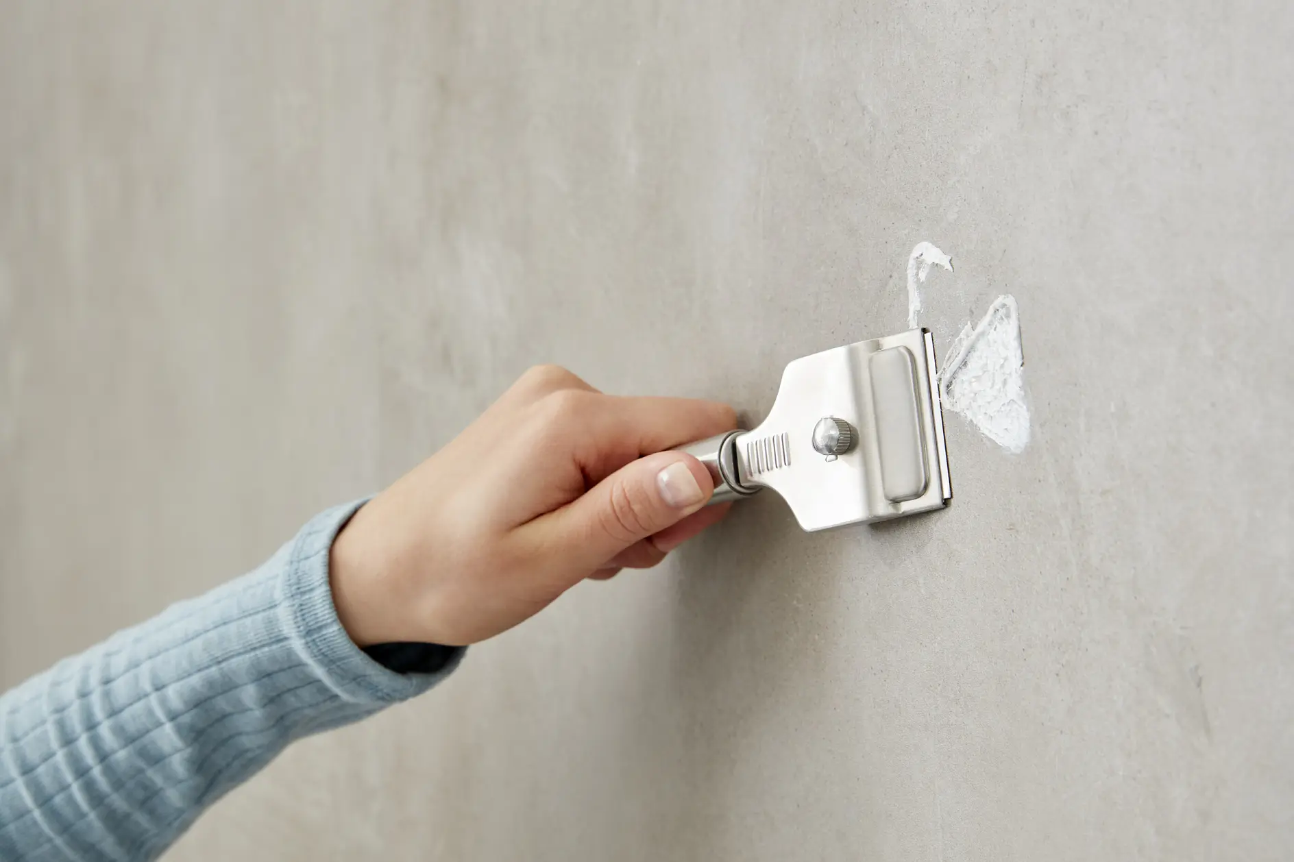 A person in a blue long-sleeve shirt uses a handheld scraper to remove white paint from a gray wall. The image focuses on the hand gripping the scraper, with the paint being chipped away using tesa tape. (This text has been generated by AI)