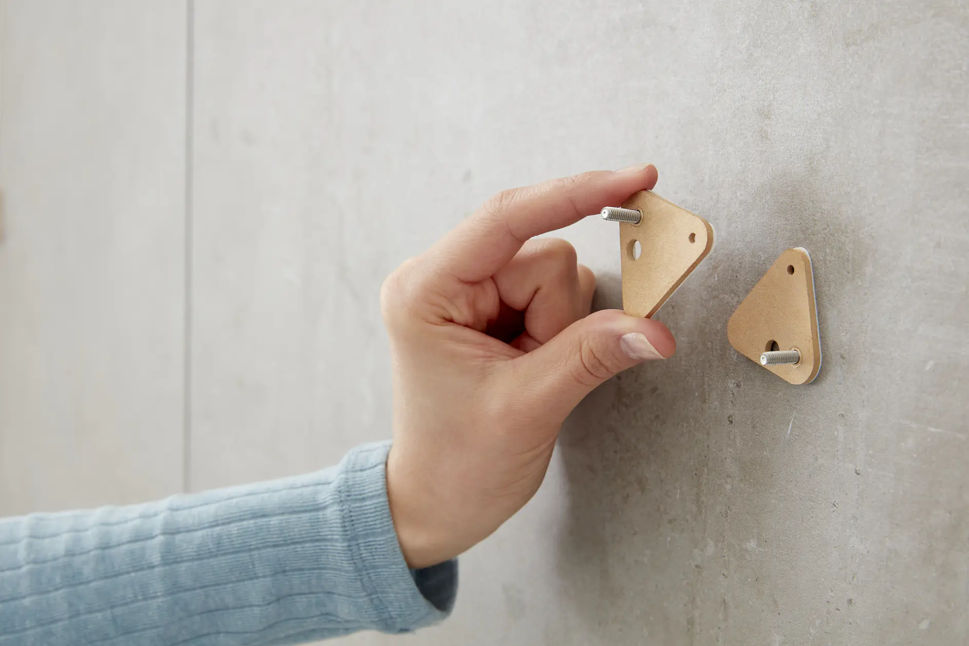 A hand in a blue sleeve attaches a triangular wooden object with a metal peg onto a gray wall using tesa tape. Another identical piece with a peg is already fixed on the wall. (This text has been generated by AI)