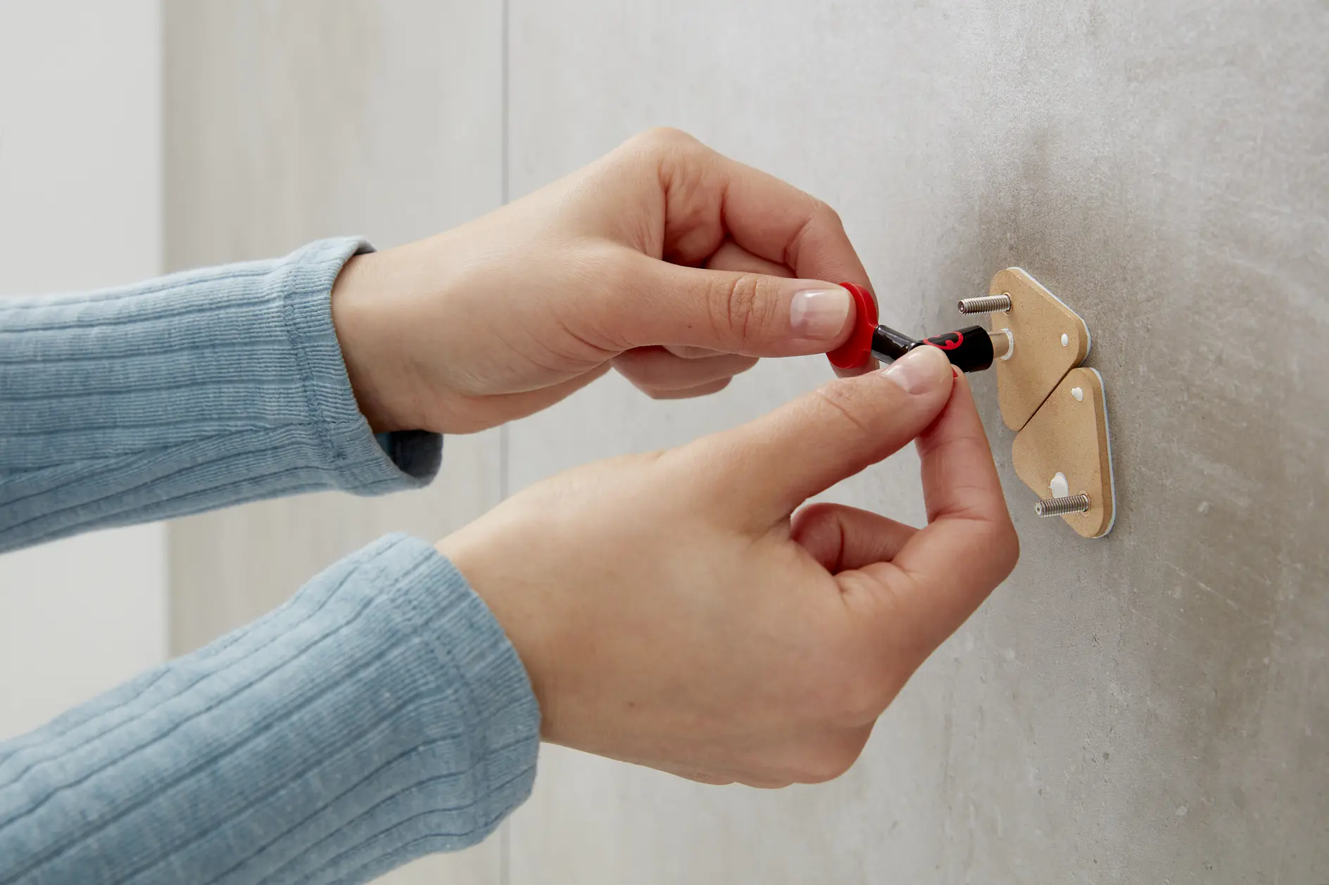 A person in a light blue sweater is using a red-handled screwdriver to tighten a screw on a wall bracket with two hooks, secured with tesa tape. The wall has a smooth, light-colored surface. (This text has been generated by AI)