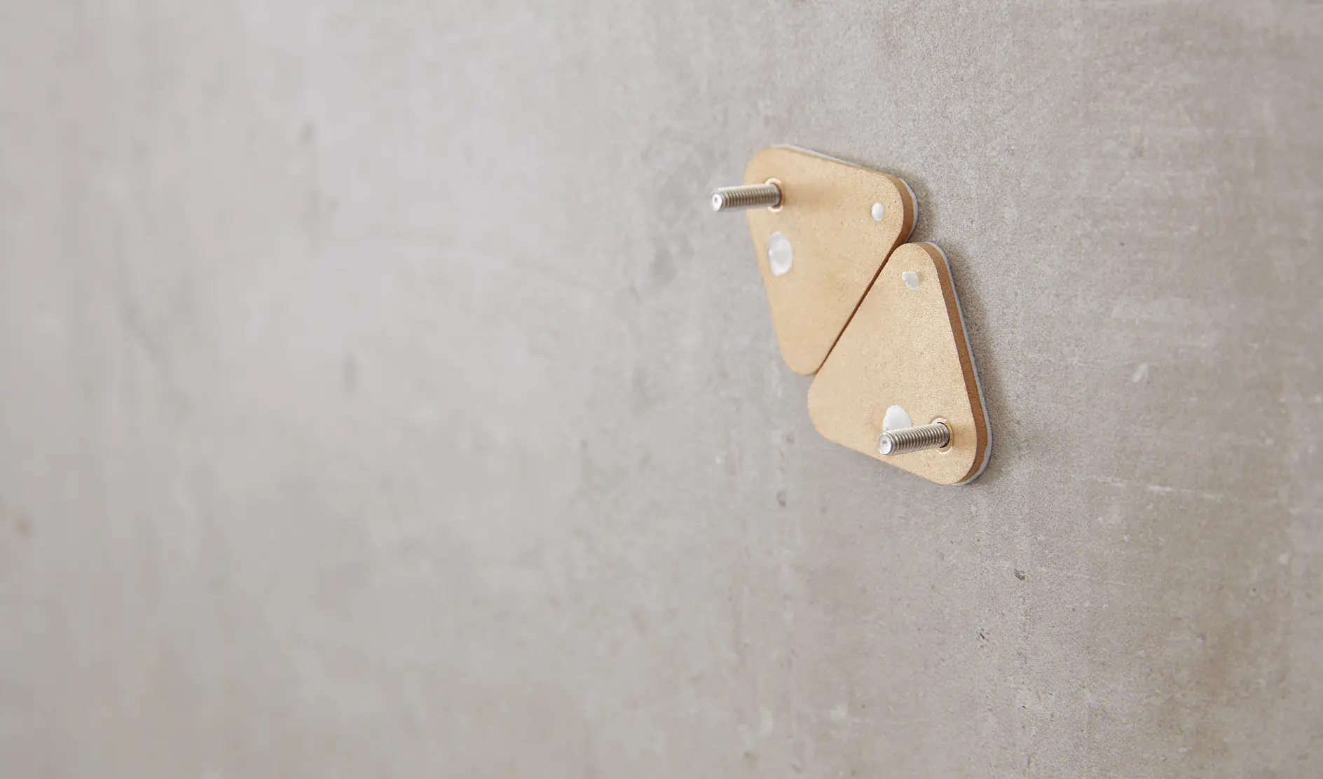 Two triangular cardboard pieces with metal pegs are mounted on a smooth, gray wall using tesa tape. The objects are positioned closely together near the center of the image. The background is plain and unadorned. (This text has been generated by AI)