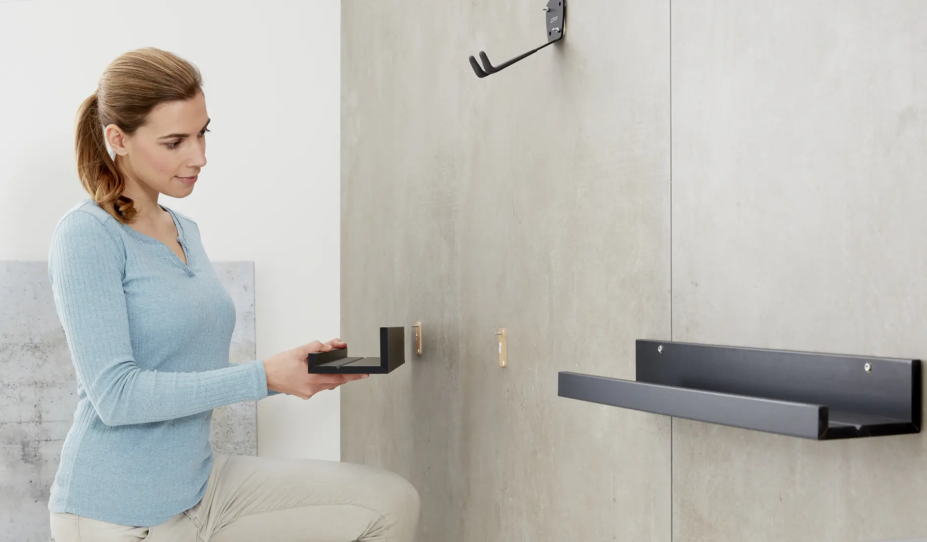 A woman in a light blue sweater kneels on the floor, holding a black metal wall bracket and facing a gray wall. Another identical bracket is mounted on the wall, with a black hook fixed above it using tesa tape. (This text has been generated by AI)