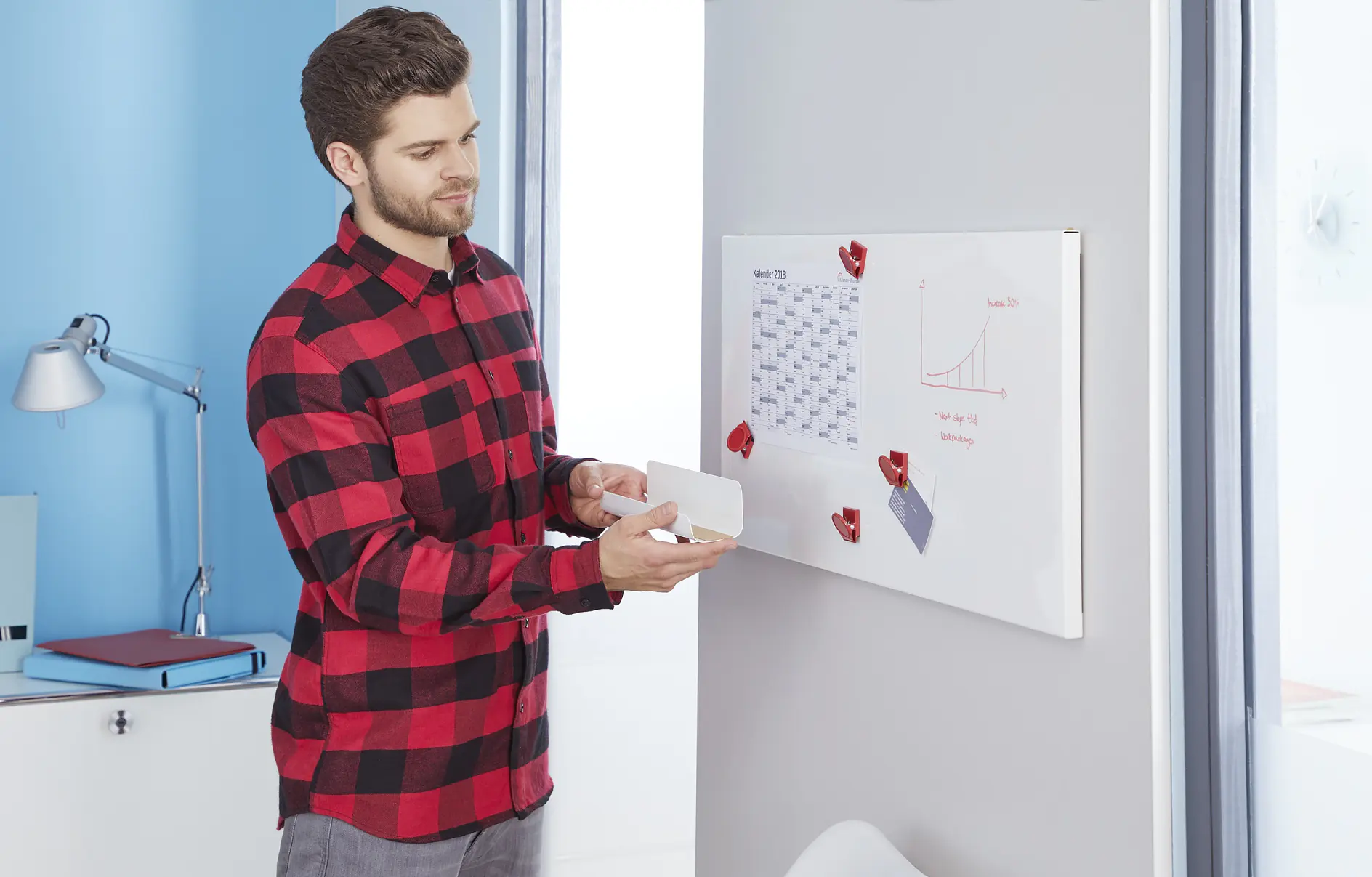 A man in a red and black checkered shirt stands by a whiteboard. The board displays a calendar and a graph, with red magnets and tesa tape holding papers. He holds a marker or eraser, appearing to be preparing or reviewing content. A desk and lamp are in the background. (This text has been generated by AI)