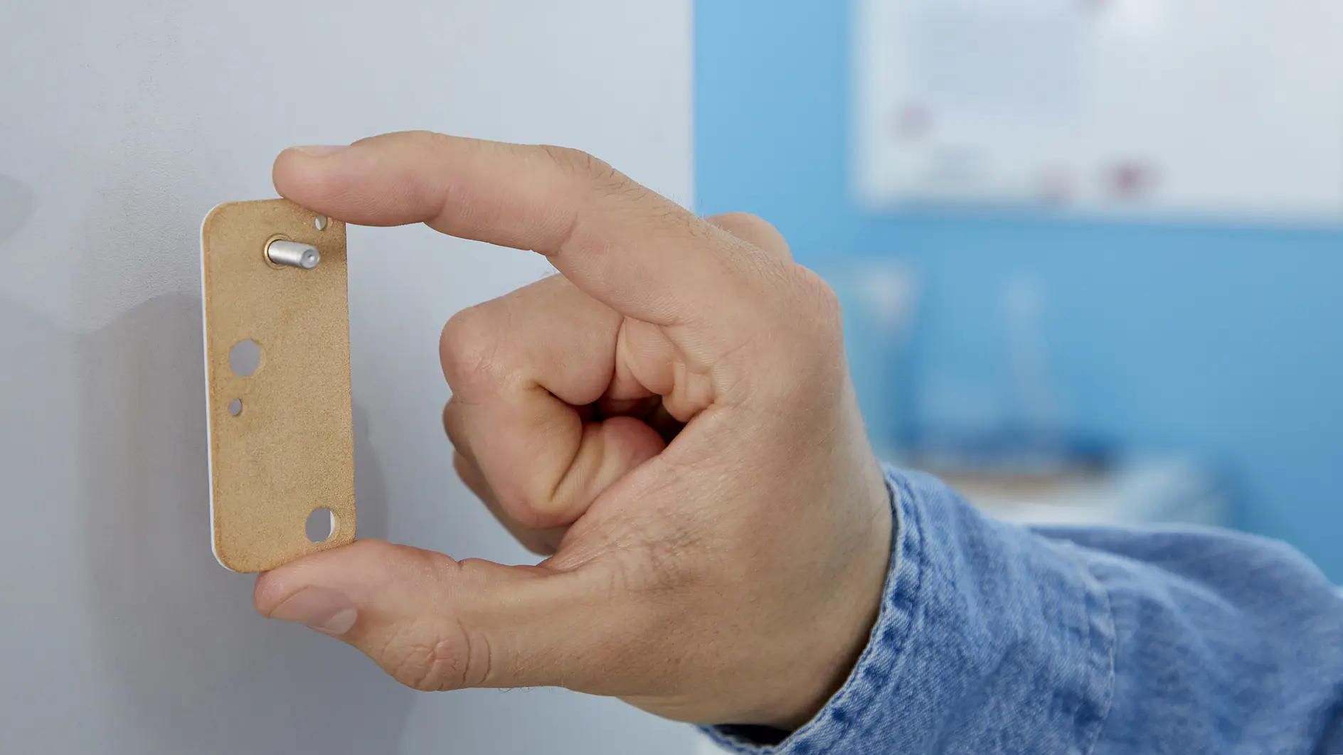 A hand in a blue denim sleeve is holding a rectangular piece of light brown material with a small metal pin. The hand is pressing it against a vertical surface, secured with tesa tape. A blurry whiteboard is visible in the background. (This text has been generated by AI)