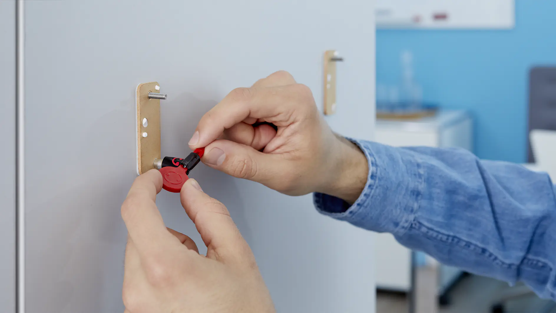 A person in a blue denim shirt adjusts a red and black lockout device on a cabinet door handle in an office environment using tesa tape. The door handle is mounted on a light-colored surface. (This text has been generated by AI)