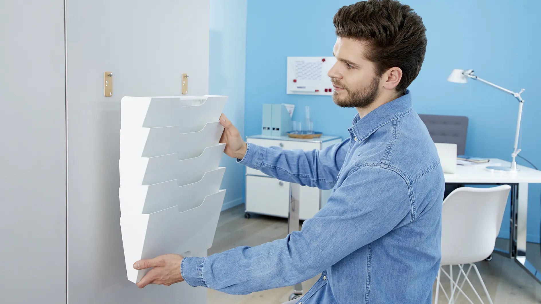 A man in a blue denim shirt is attaching a wall-mounted file organizer on a white wall in a modern office space using tesa tape. The room features a light blue wall, white furniture, and a desk lamp in the background. (This text has been generated by AI)