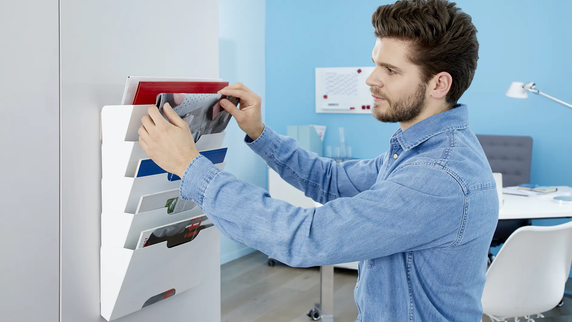 A man in a denim shirt is organizing folders in a wall-mounted file holder using tesa tape. He is in a brightly lit office with blue walls, a desk lamp, and office supplies in the background. (This text has been generated by AI)
