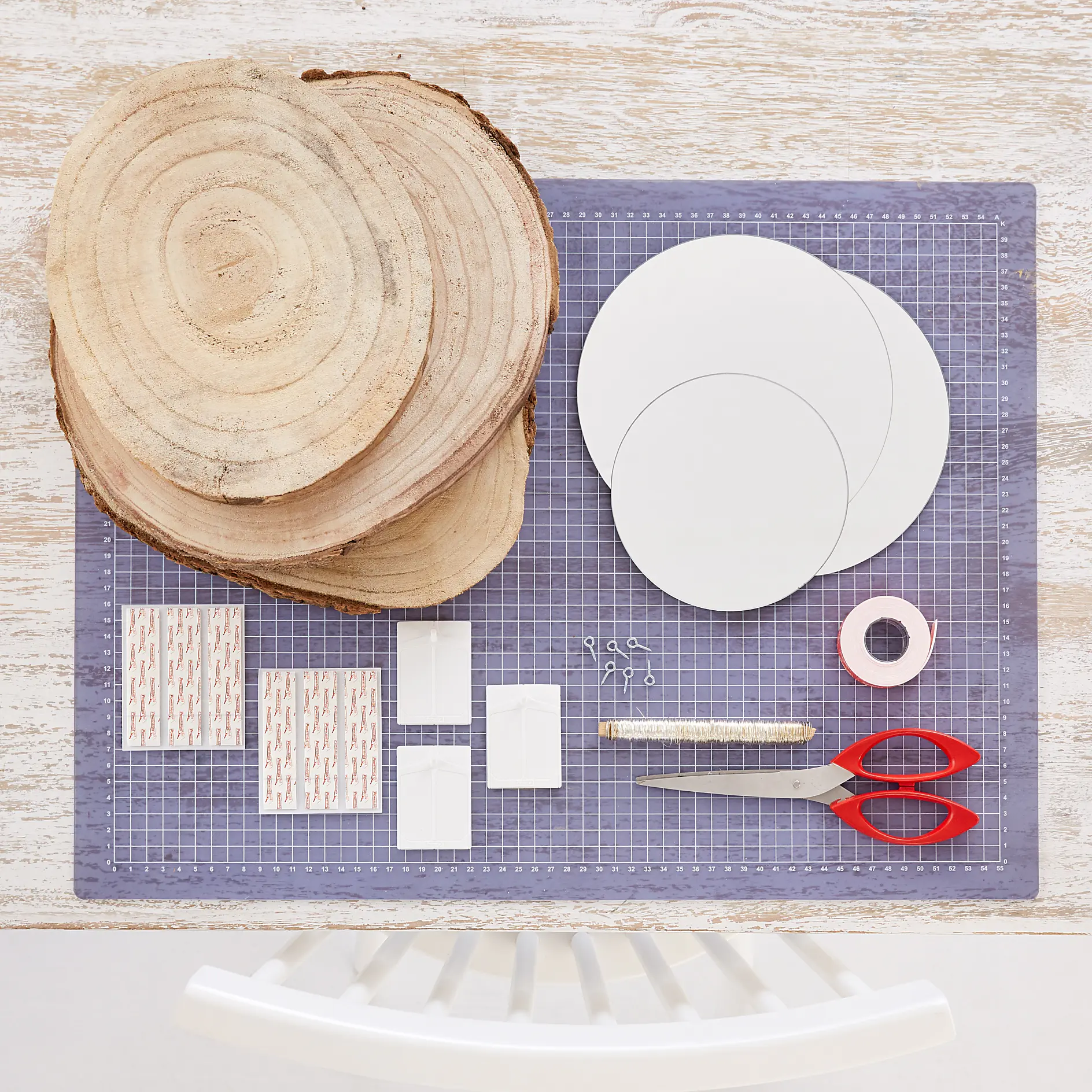 A crafting setup with wooden slices, round white papers, tesa tape strips, string, a paper roll, scissors, and a wire cutter on a cutting mat. A white chair is partially visible at the bottom. (This text has been generated by AI)