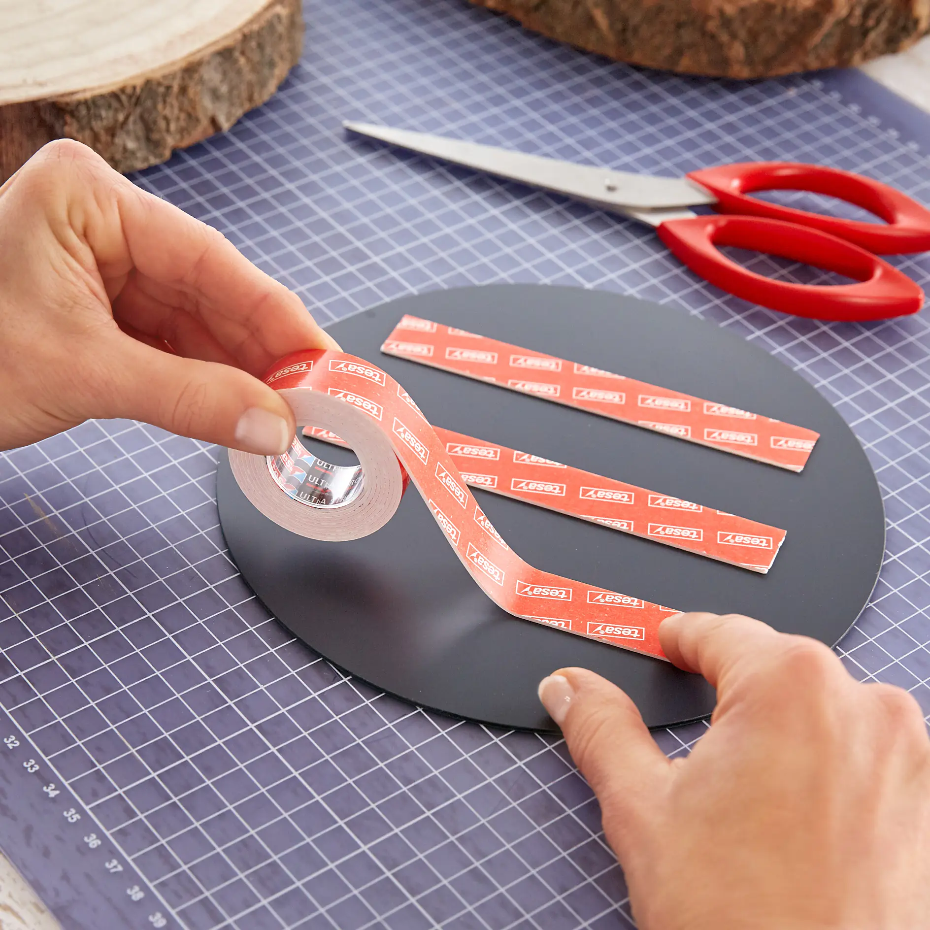A person is applying strips of red tesa tape onto a round black surface. A pair of red scissors and a grid-patterned cutting mat are in the background. (This text has been generated by AI)