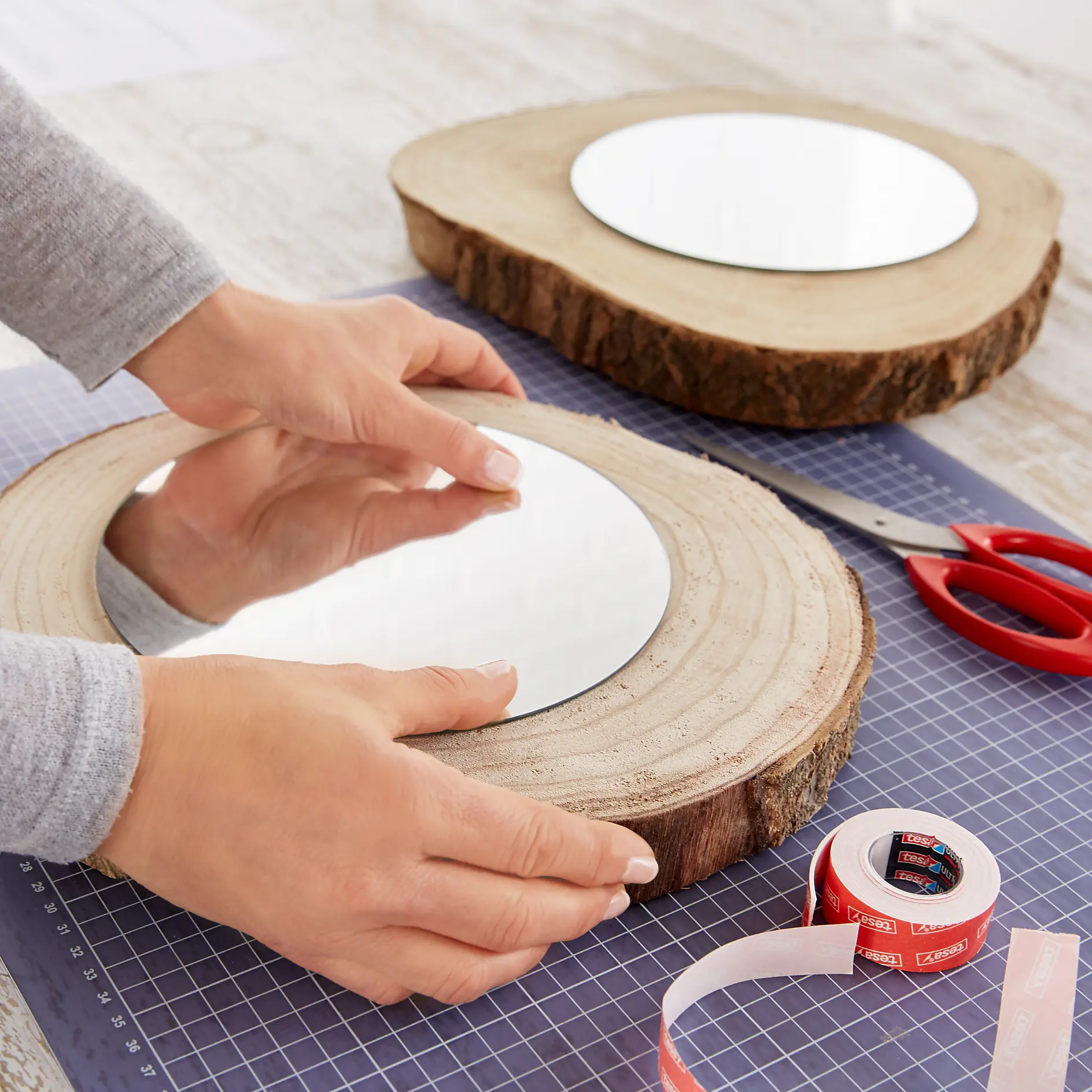 Close-up of a person placing a round mirror on a wood slice. Another similar mirror is nearby. The scene includes a cutting mat, red scissors, measuring tape, and tesa tape on a wooden surface. (This text has been generated by AI)