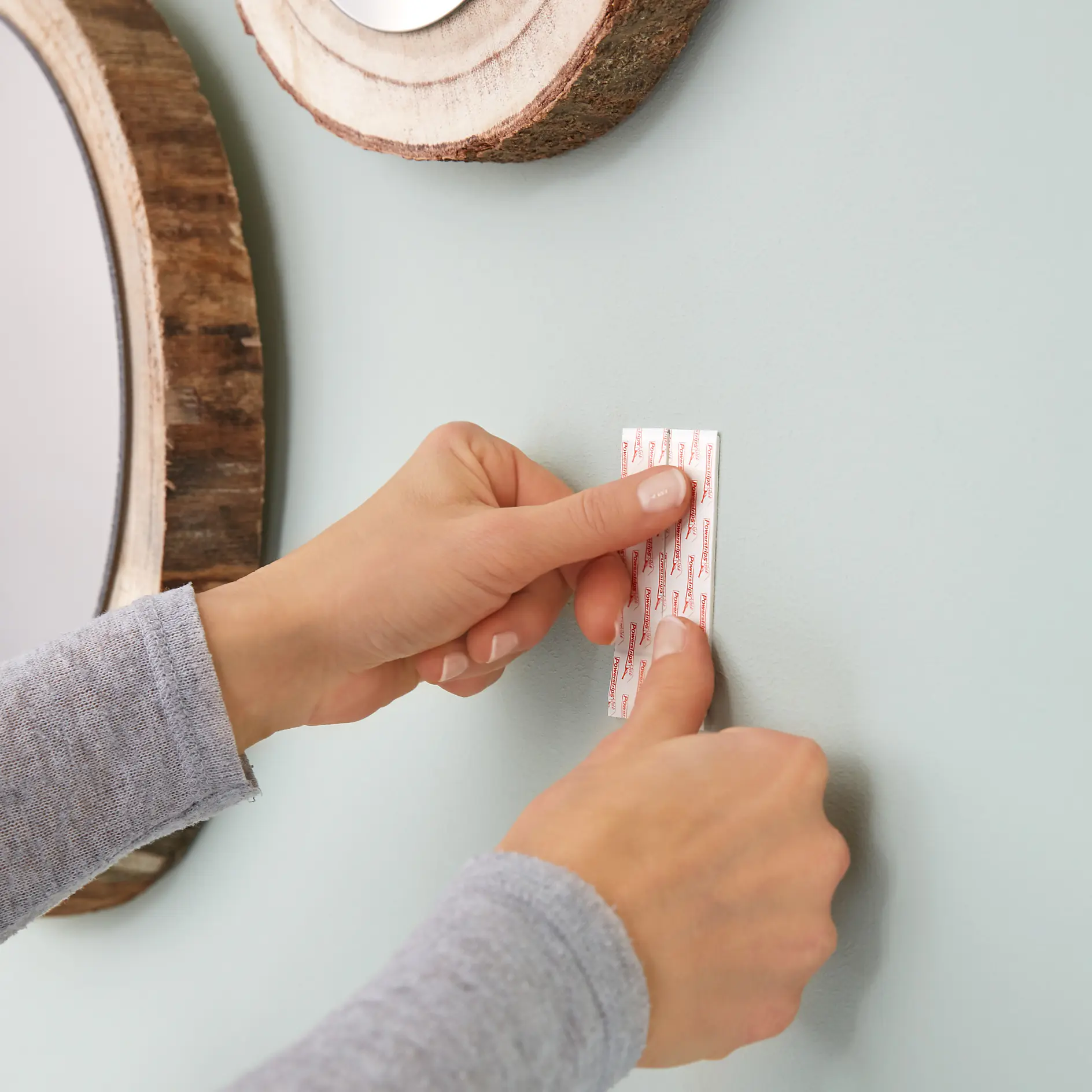 Two hands affix a white strip of tesa tape to a light green wall. Wooden round wall decorations are partially visible. The person wears a long-sleeved gray shirt. (This text has been generated by AI)