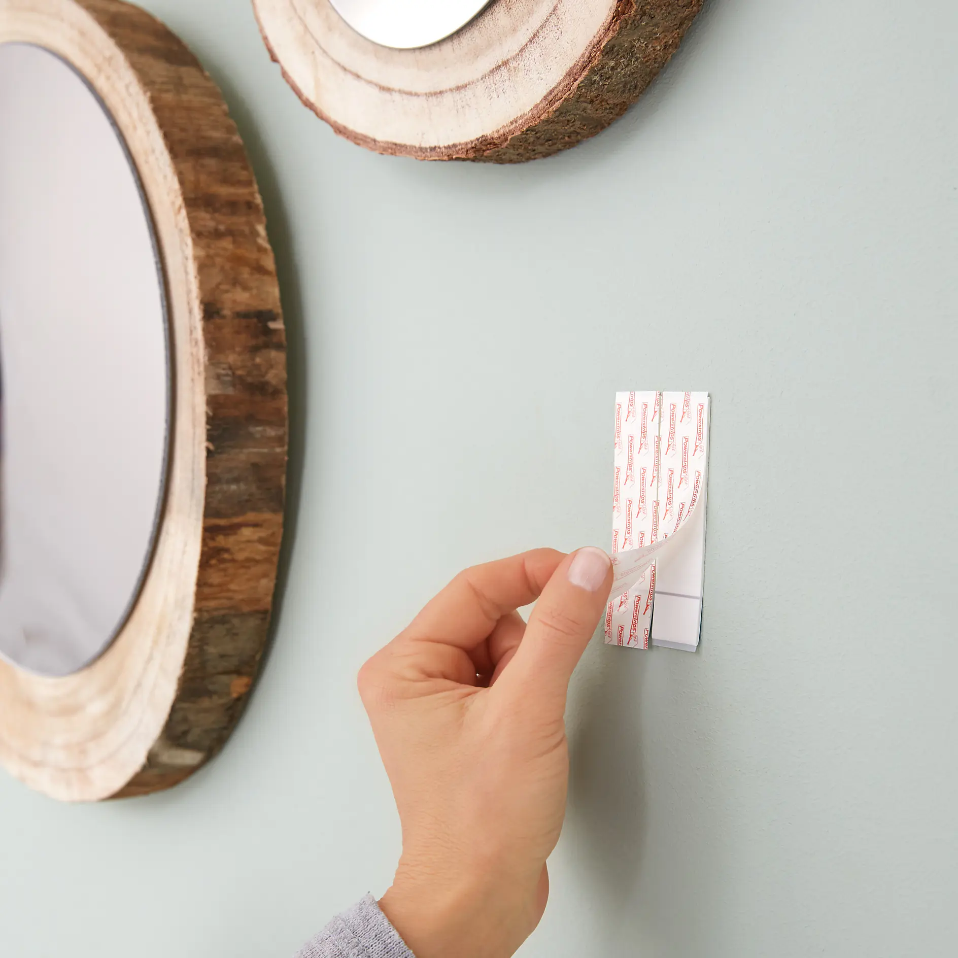 A hand is peeling off a tesa tape adhesive strip on a wall. The wall is painted light green. Two round mirrors with wooden frames are visible, one partially out of the frame on the left side. (This text has been generated by AI)