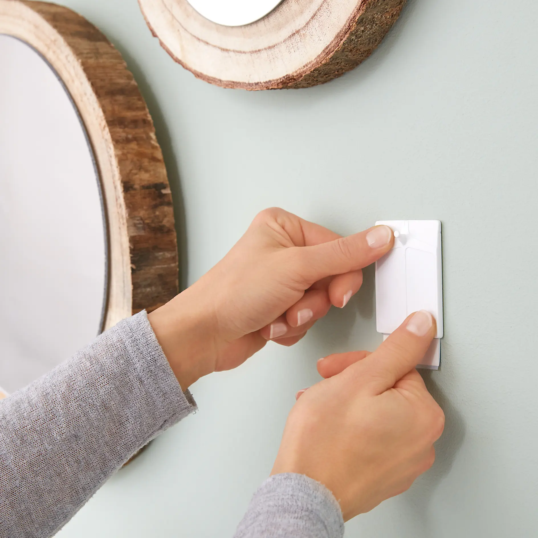 Hands applying a white tesa tape strip to a light green wall, between round wooden-framed mirrors. The person is wearing a gray long-sleeve shirt. (This text has been generated by AI)