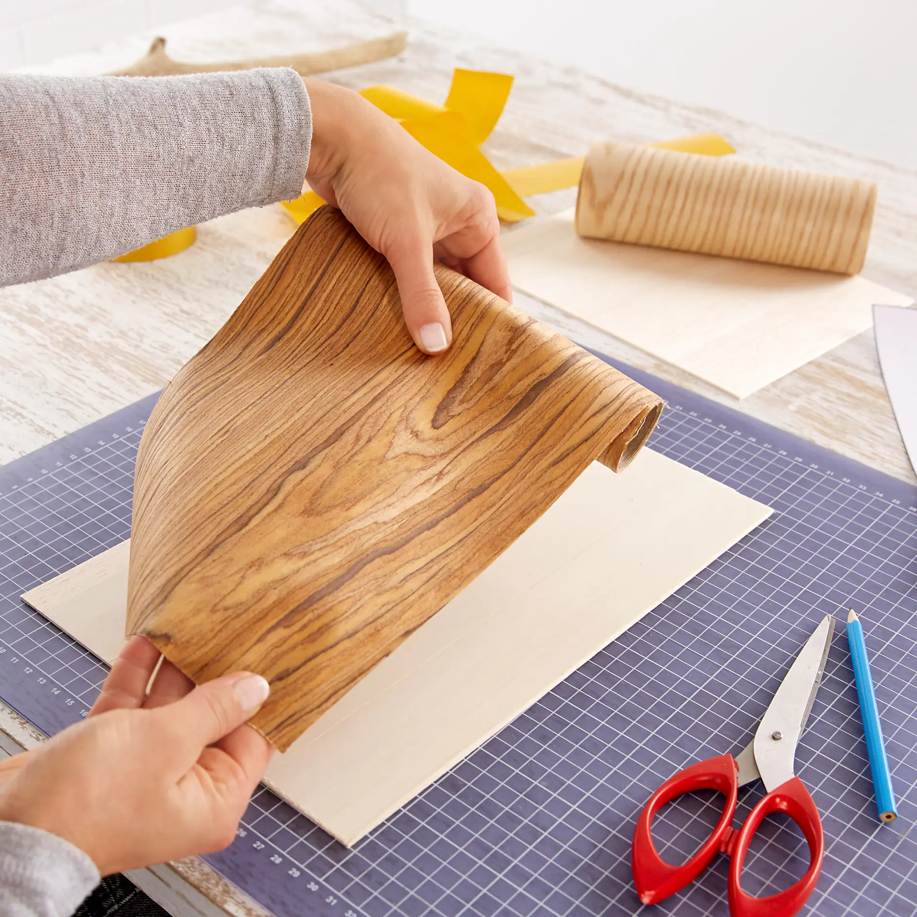 Two hands are holding a sheet of wood-patterned tesa tape above a piece of wood on a cutting mat. Nearby are a pair of red scissors, a blue pencil, and a black knife on the table. Other rolls of tesa tape are also visible. (This text has been generated by AI)