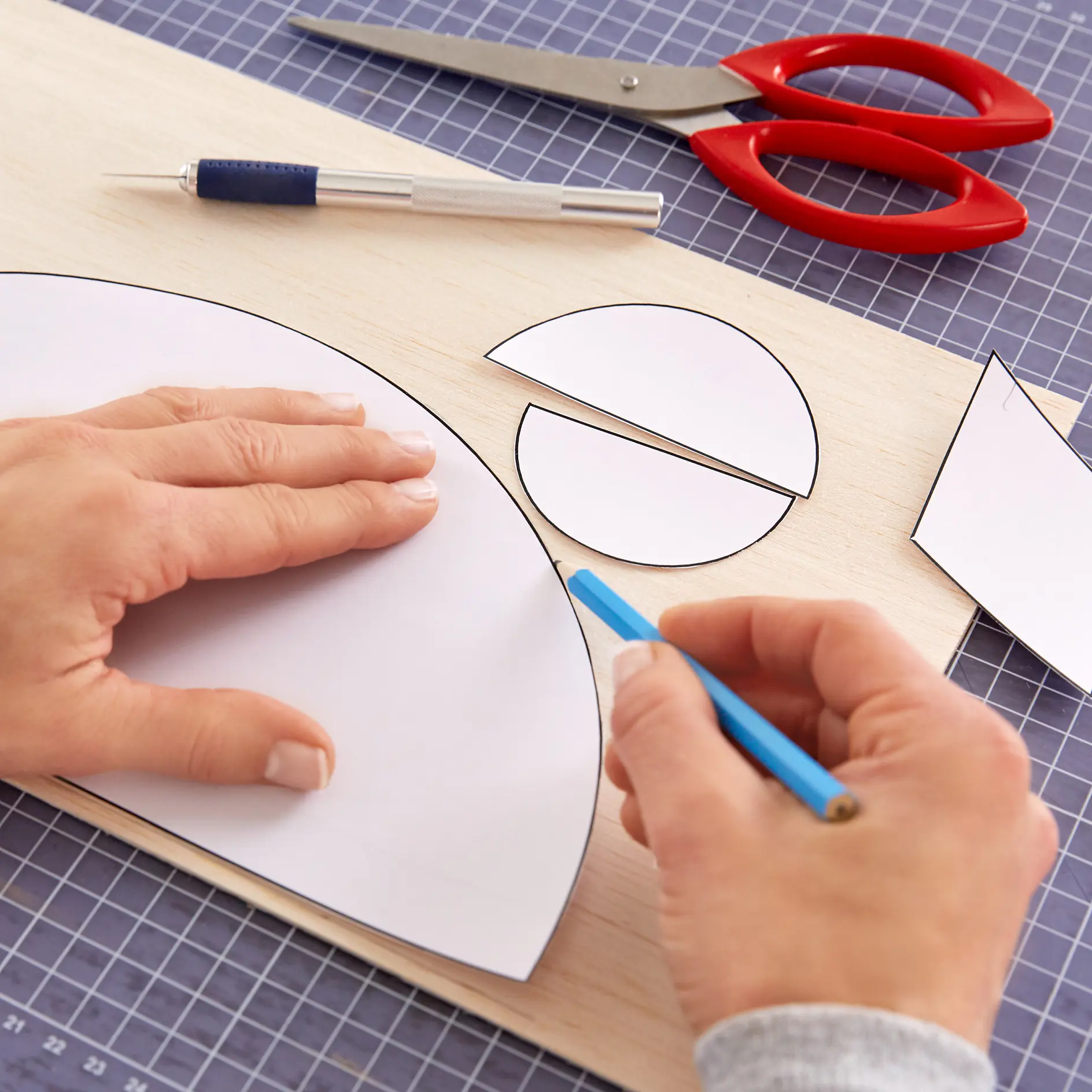 Hands trace circular and semicircular shapes onto white paper placed on a wooden board. A blue pencil is used for tracing. Red scissors and a craft knife are on a grid-patterned cutting mat, along with tesa tape for securing the paper during the process. (This text has been generated by AI)