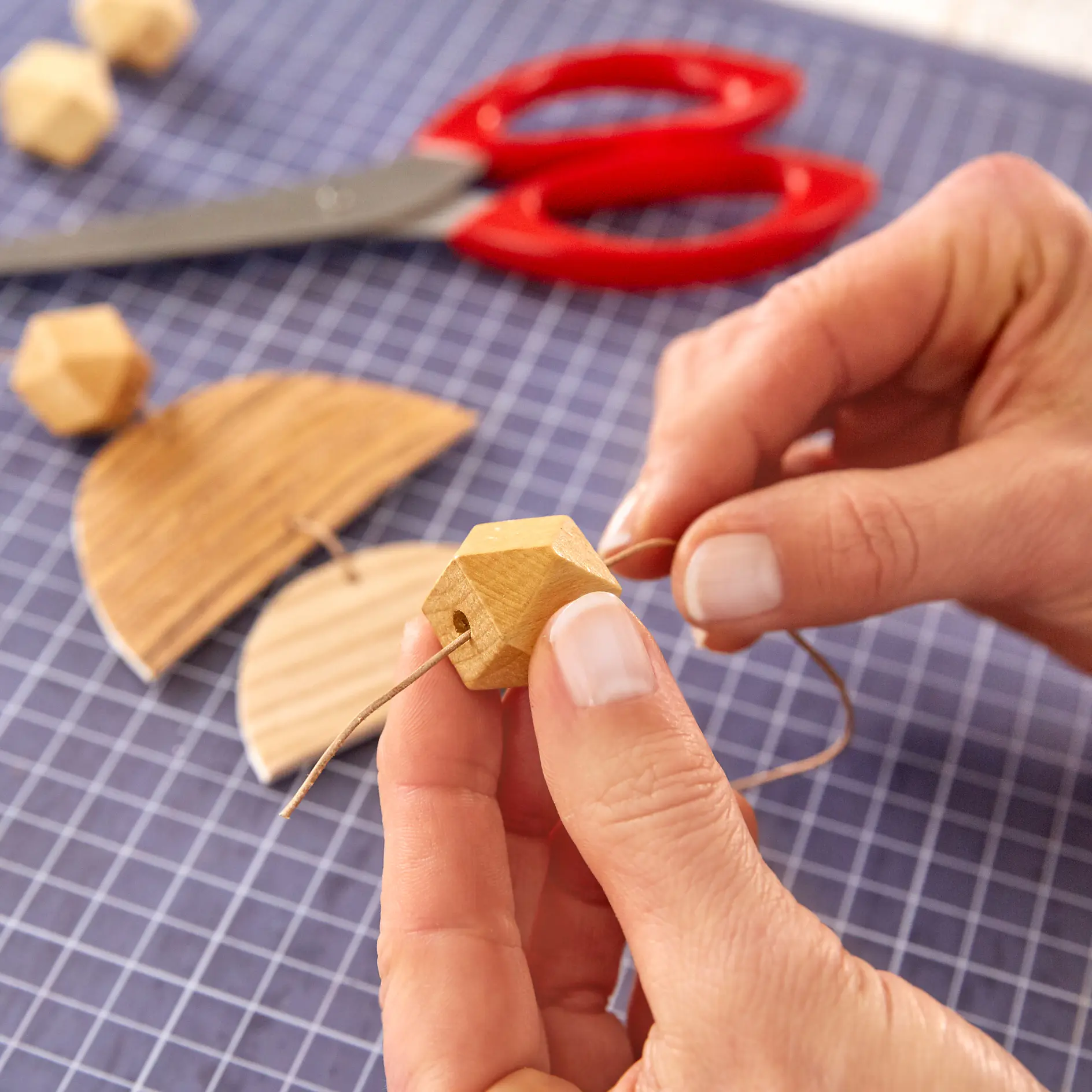 Hands are threading a wooden bead onto a string. A pair of red scissors and wooden shapes are in the background on a grid-patterned surface, along with a roll of tesa tape. (This text has been generated by AI)