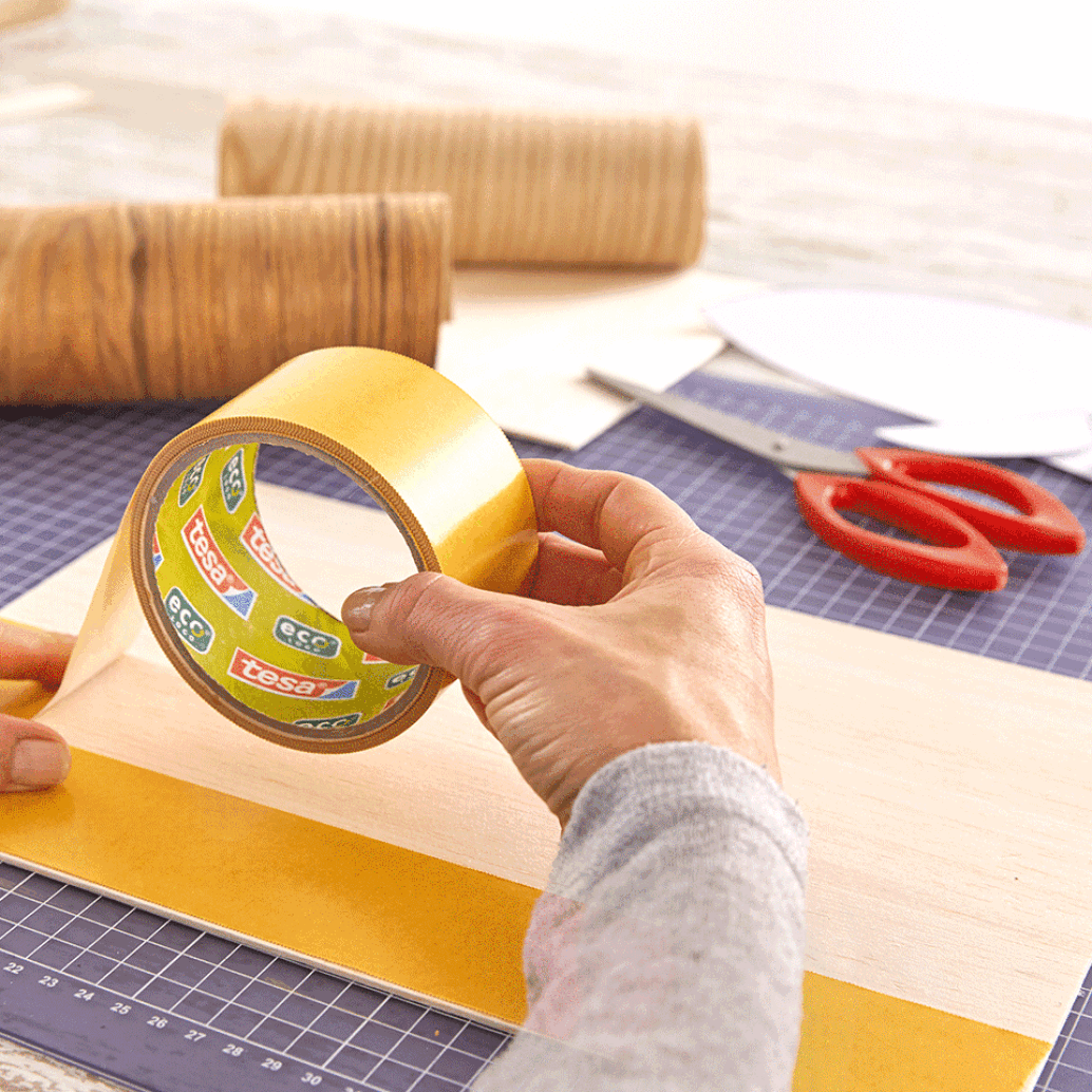 A person holding a roll of yellow tesa tape is applying it to a piece of wood. Red scissors and two rolls of brown paper are in the background, all placed on a grid-patterned cutting mat. (This text has been generated by AI)