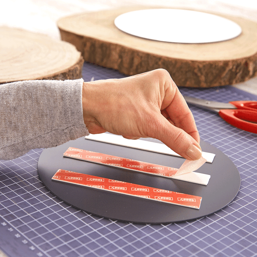 A person peels the backing off adhesive strips on a circular black object, placed on a cutting mat. Scissors and a piece of round wood are nearby, alongside tesa tape. (This text has been generated by AI)