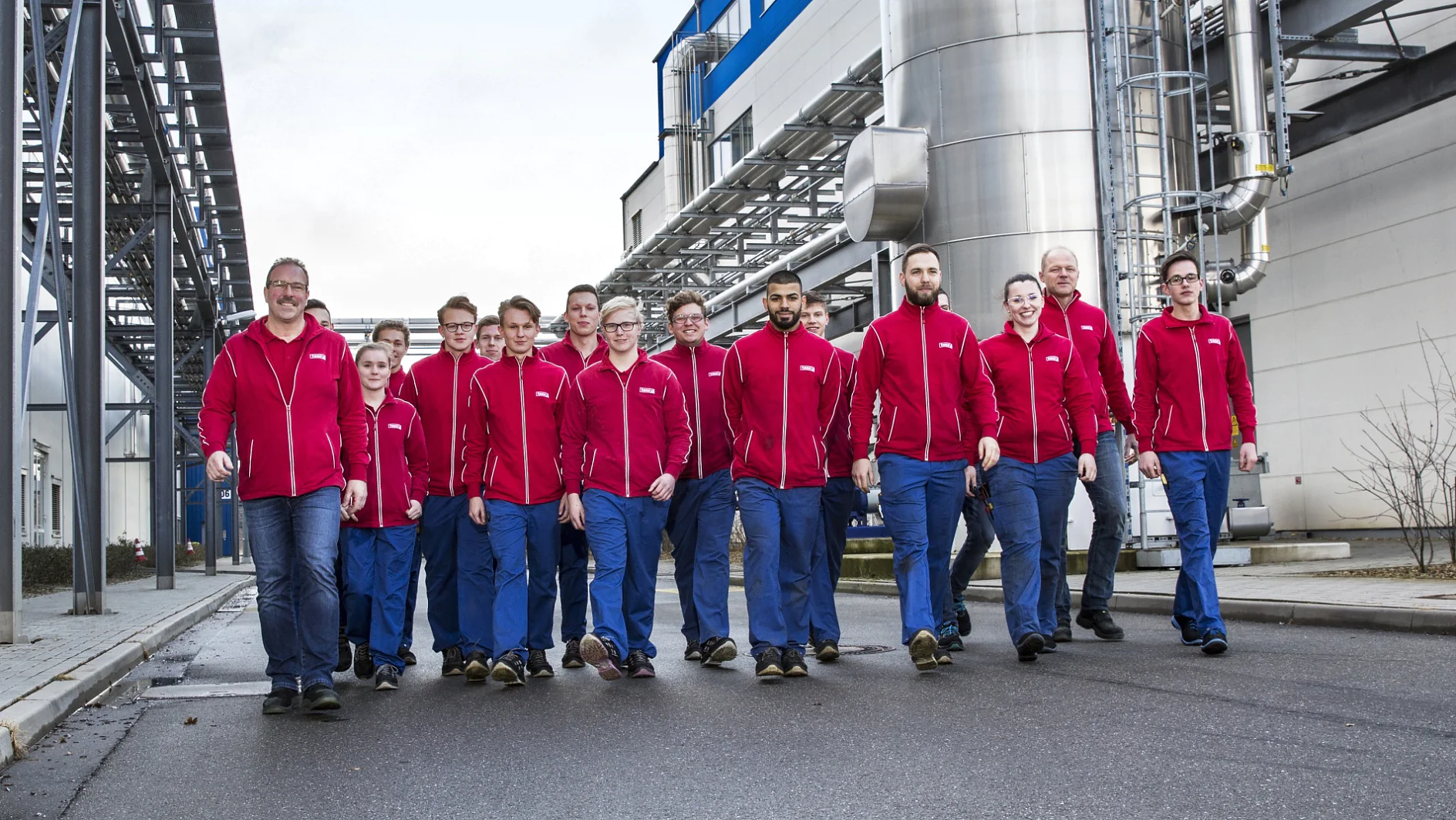 Eine Gruppe von vierzehn Personen in roten Jacken und blauen Hosen geht eine gepflasterte Straße zwischen Industriegebäuden entlang. Sie scheinen sich in einer Fabrik zu befinden, im Hintergrund sind Metallkonstruktionen und Tesa-Klebeband zu sehen. (This text has been generated by AI)