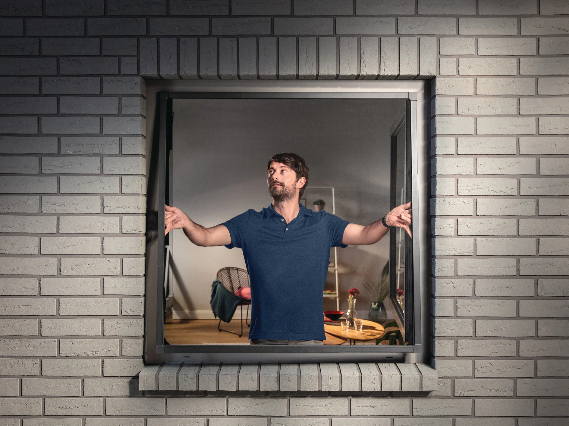 A man in a blue shirt is looking out of a window at night. He is standing inside a room with light-colored brick walls, and the window is open. Behind him, a table with a red flower and a partially visible chair can be seen. (This text has been generated by AI)