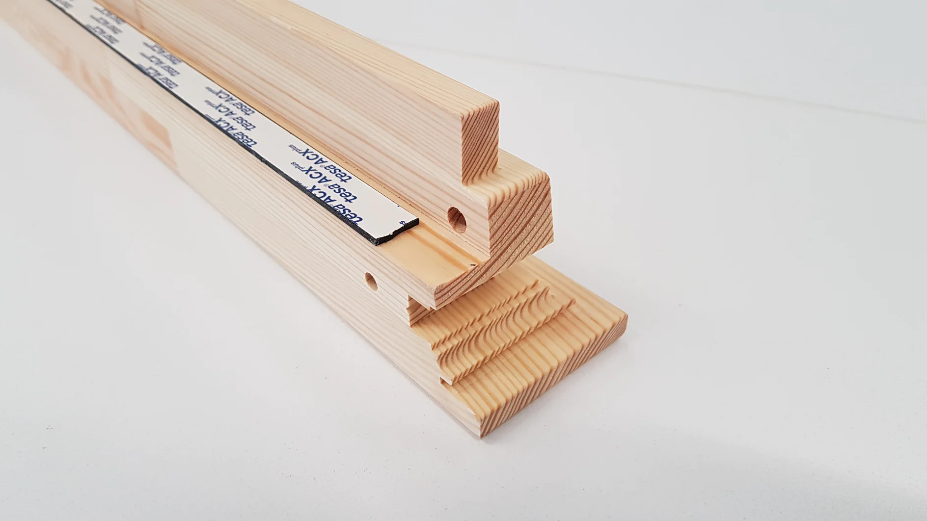 Ein Stapel Holzbretter mit präzisen Schnitten und Löchern. Das Holz scheint hell zu sein, mit sichtbaren Maserungen. An einem der Bretter ist teilweise ein Streifen Tesa-Klebeband befestigt. (This text has been generated by AI)