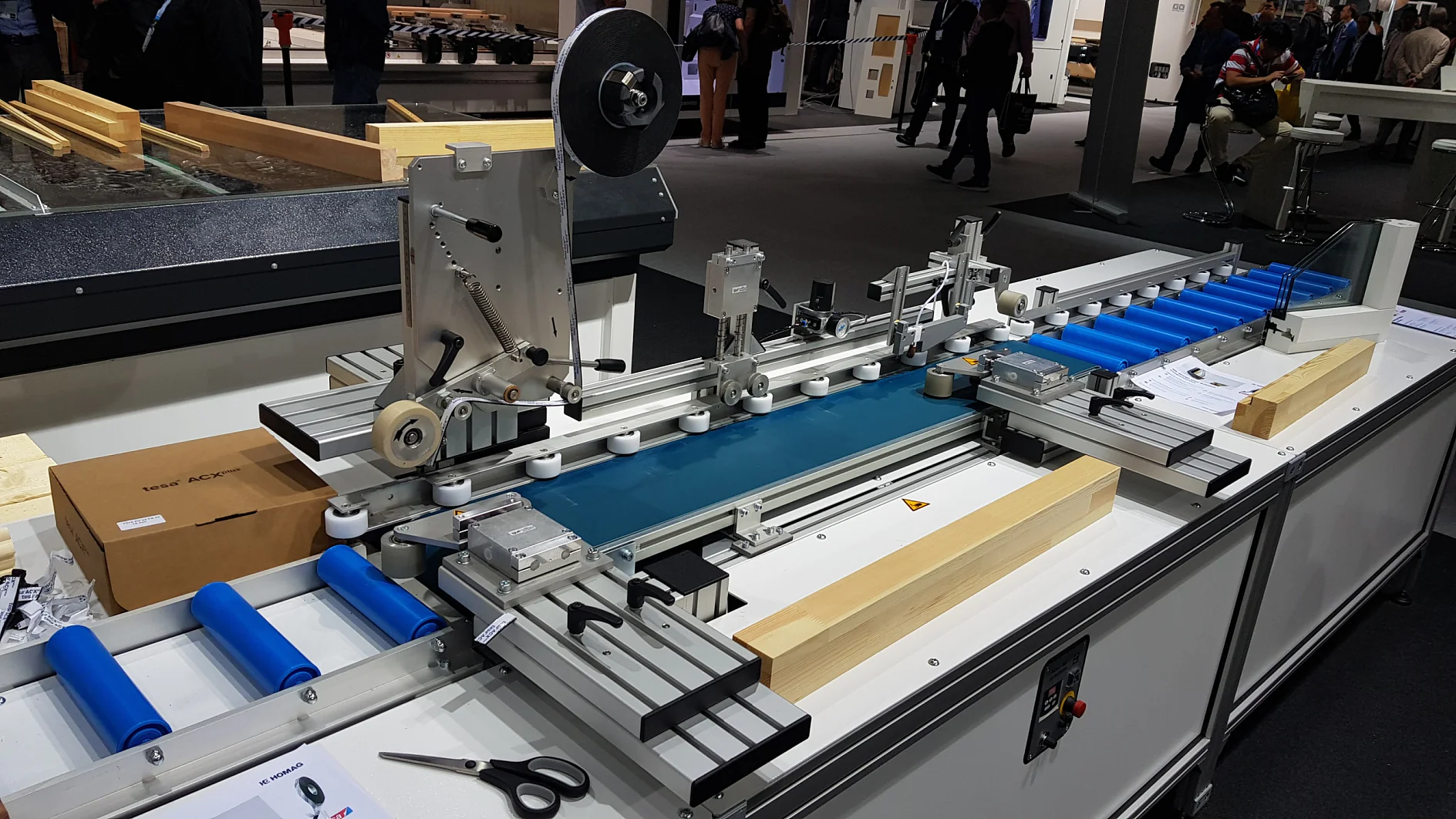 In einer industriellen Umgebung wird eine komplexe Maschine mit Förderband, Walzen und mechanischen Komponenten gezeigt. Die Maschine verarbeitet flache Materialien. In der Nähe sind Holzbretter und eine geschlossene Schere sowie Tesa-Klebeband zu sehen. Im Hintergrund sind Menschen zu sehen. (This text has been generated by AI)