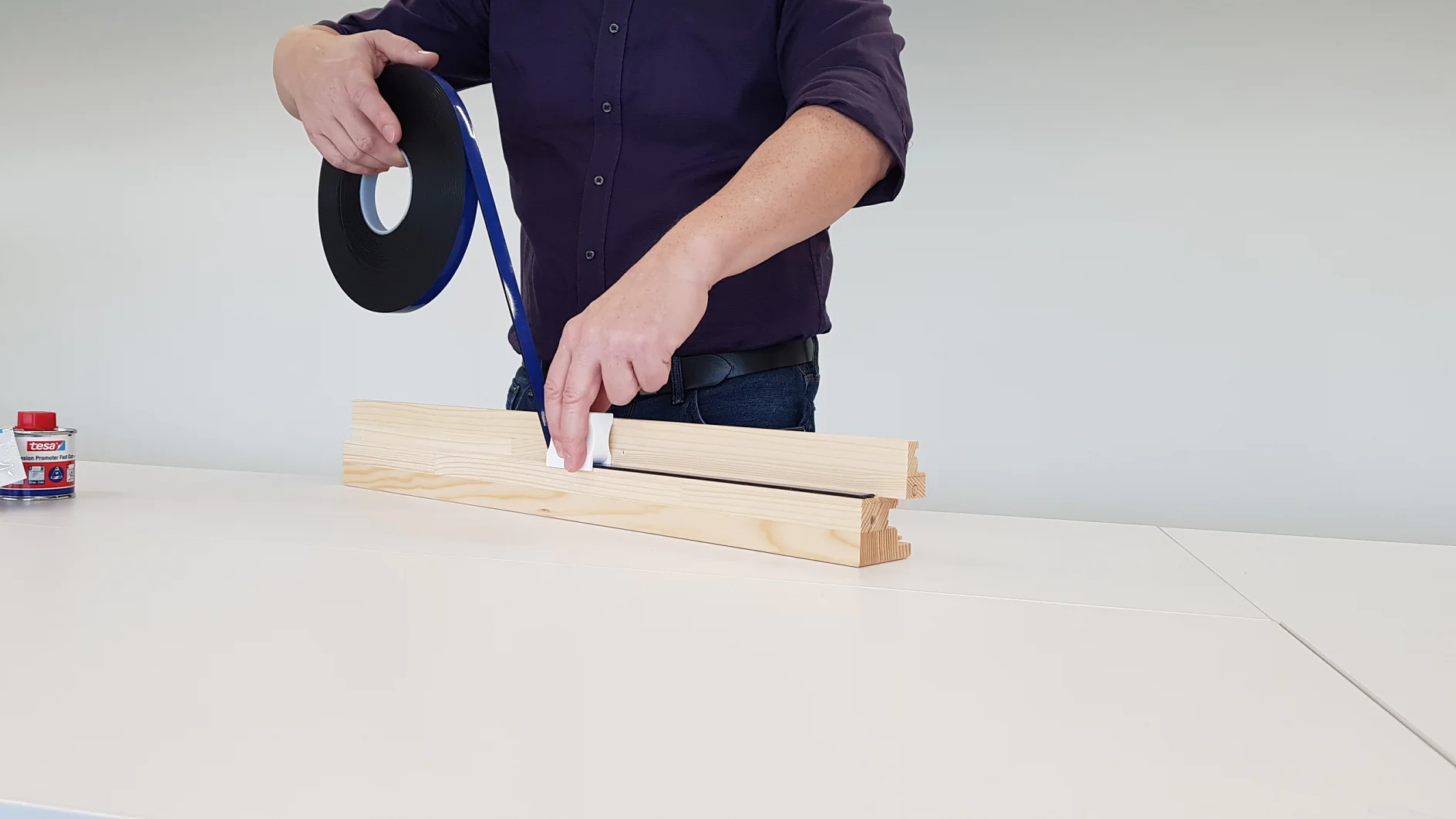 Eine Person im dunklen Hemd klebt schwarzes Tesa-Klebeband auf ein Stück helles Holz auf einem weißen Tisch. Links auf dem Tisch steht ein kleiner roter Behälter mit weißem Etikett. (This text has been generated by AI)