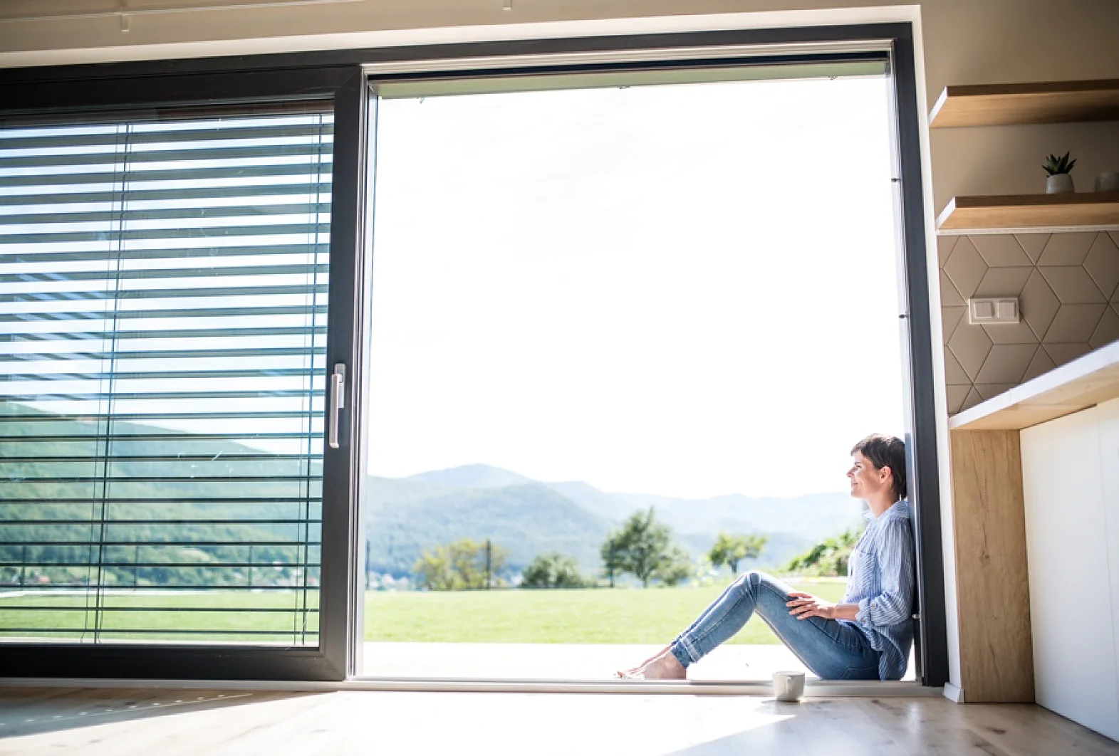 Eine Person sitzt am Rand einer offenen Glasschiebetür und blickt nach draußen. Sie sitzt mit gekreuzten Beinen auf dem Boden. Daneben steht ein Becher. Der Blick zeigt eine Grasfläche und Hügel unter einem hellen Himmel. Innen sind teilweise Regale sichtbar, auf denen verschiedene Gegenstände ordentlich angeordnet und mit Tesa-Klebeband befestigt sind, damit alles an seinem Platz bleibt. (This text has been generated by AI)