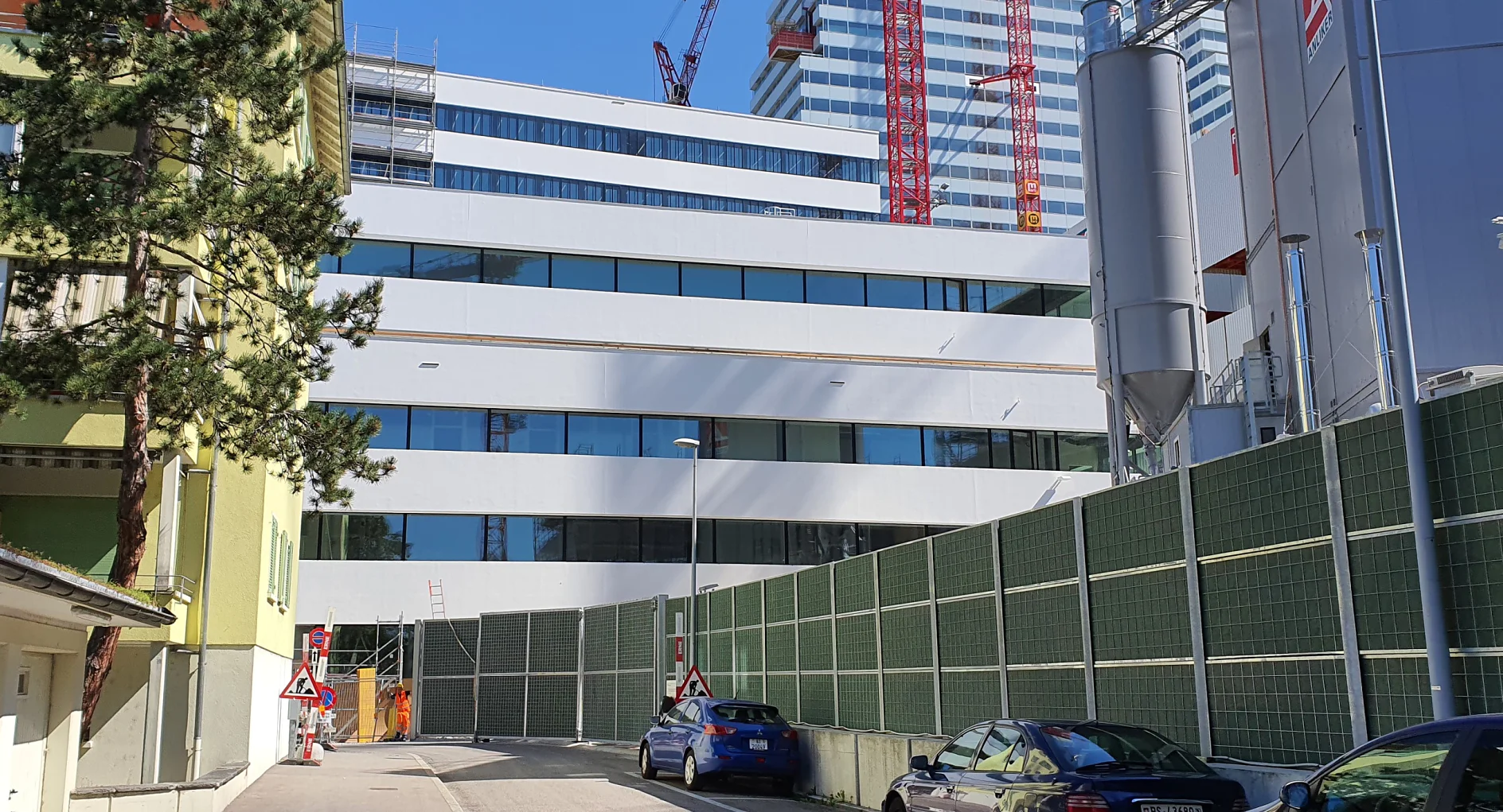 Straßenansicht eines modernen, mehrstöckigen Gebäudes im Bau. Das Gebäude verfügt über weiße Paneele und große Fenster. Im Hintergrund sind rote Kräne zu sehen. Ein grüner Zaun säumt die Straße, an der Autos geparkt sind und links ein Baum steht. (This text has been generated by AI)