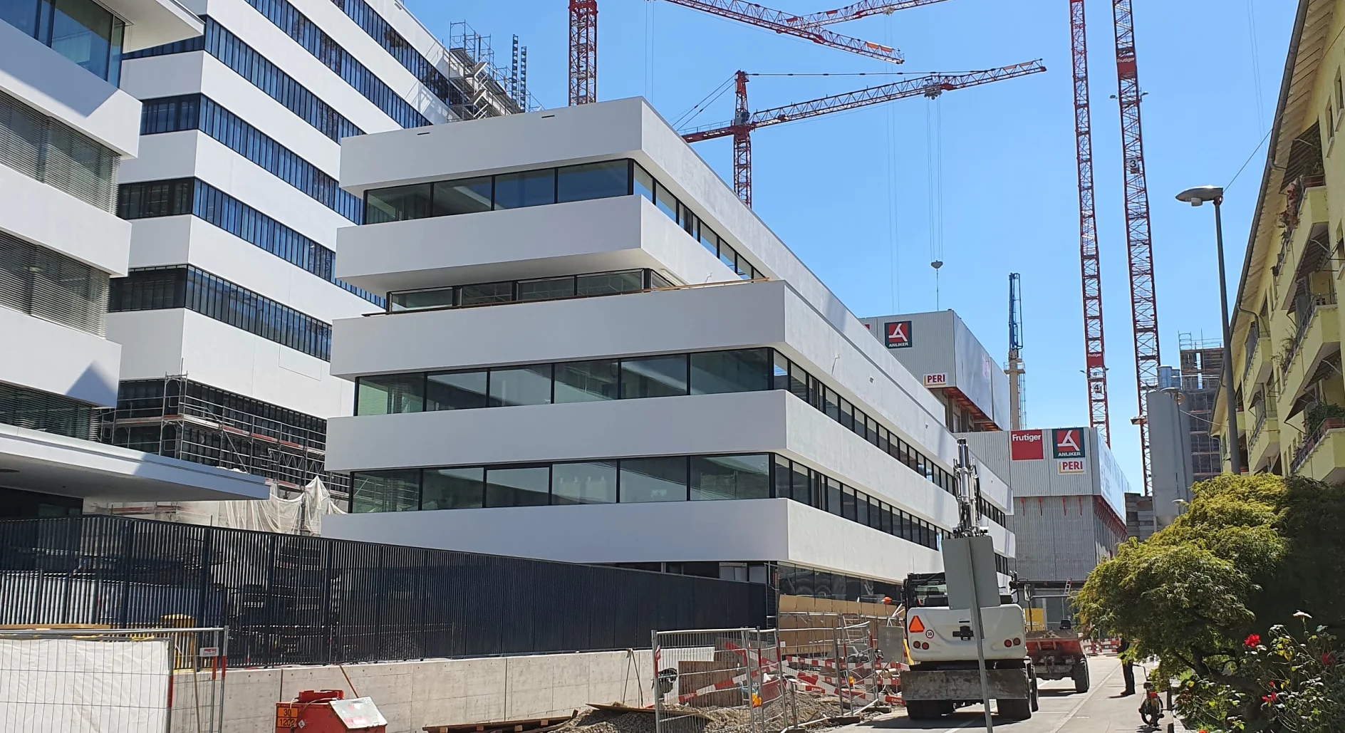 Eine Baustelle mit modernen weißen Gebäuden und großen Glasfenstern. Im Hintergrund ragen mehrere rote Kräne auf. Auf dem Boden sind Baumaterialien und Absperrungen sowie Rollen mit Tesa-Klebeband zu sehen, und im Vordergrund parkt ein weißer Lastwagen. (This text has been generated by AI)