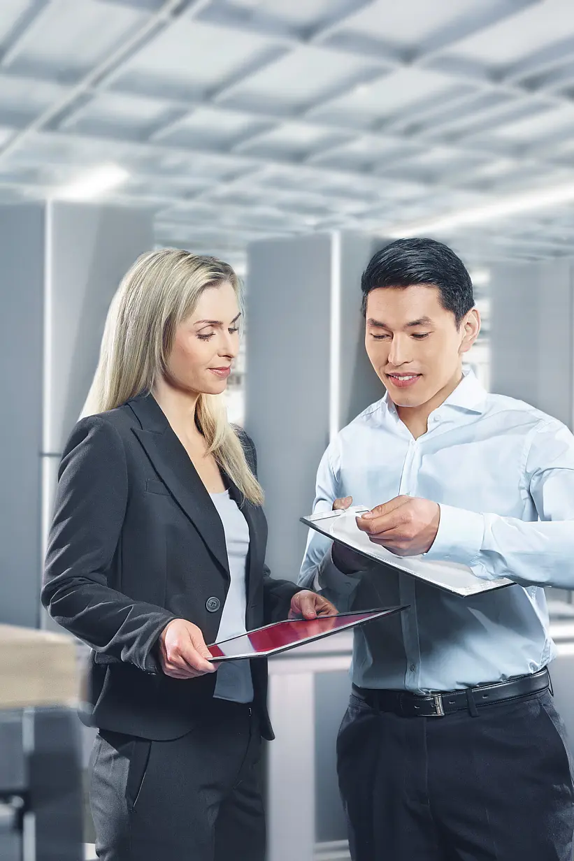 A man and a woman are discussing documents in a bright industrial setting. The woman holds a tablet, and the man holds a clipboard secured with tesa tape. They are both professionally dressed. Equipment and large metal structures are visible in the background. (This text has been generated by AI)
