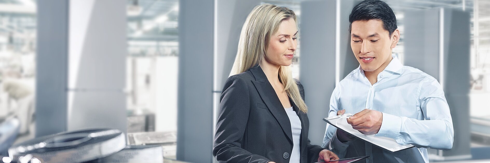 A man and a woman are discussing documents in a bright industrial setting. The woman holds a tablet, and the man holds a clipboard secured with tesa tape. They are both professionally dressed. Equipment and large metal structures are visible in the background.