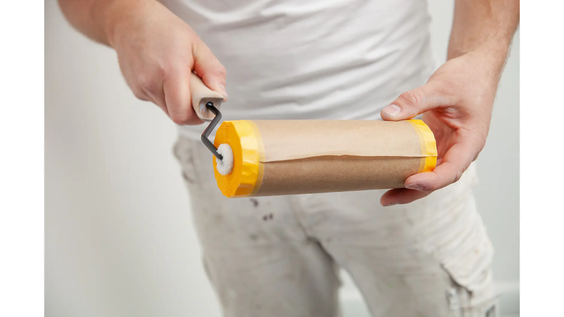 A person in a white shirt and paint-splattered pants is holding a paint roller covered with brown paper and secured at both ends with yellow tesa tape. The person appears to be preparing or cleaning the roller. (This text has been generated by AI)